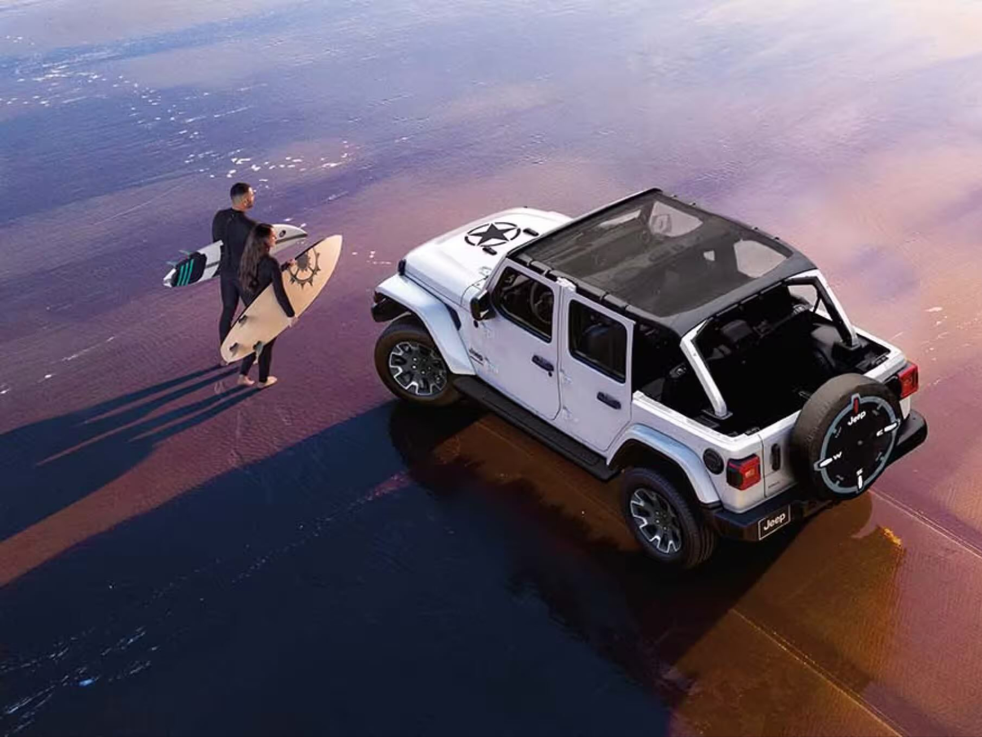 A man with a surfboard poses by a white SUV on a wet beach, with the ocean and a colorful sky in the background.