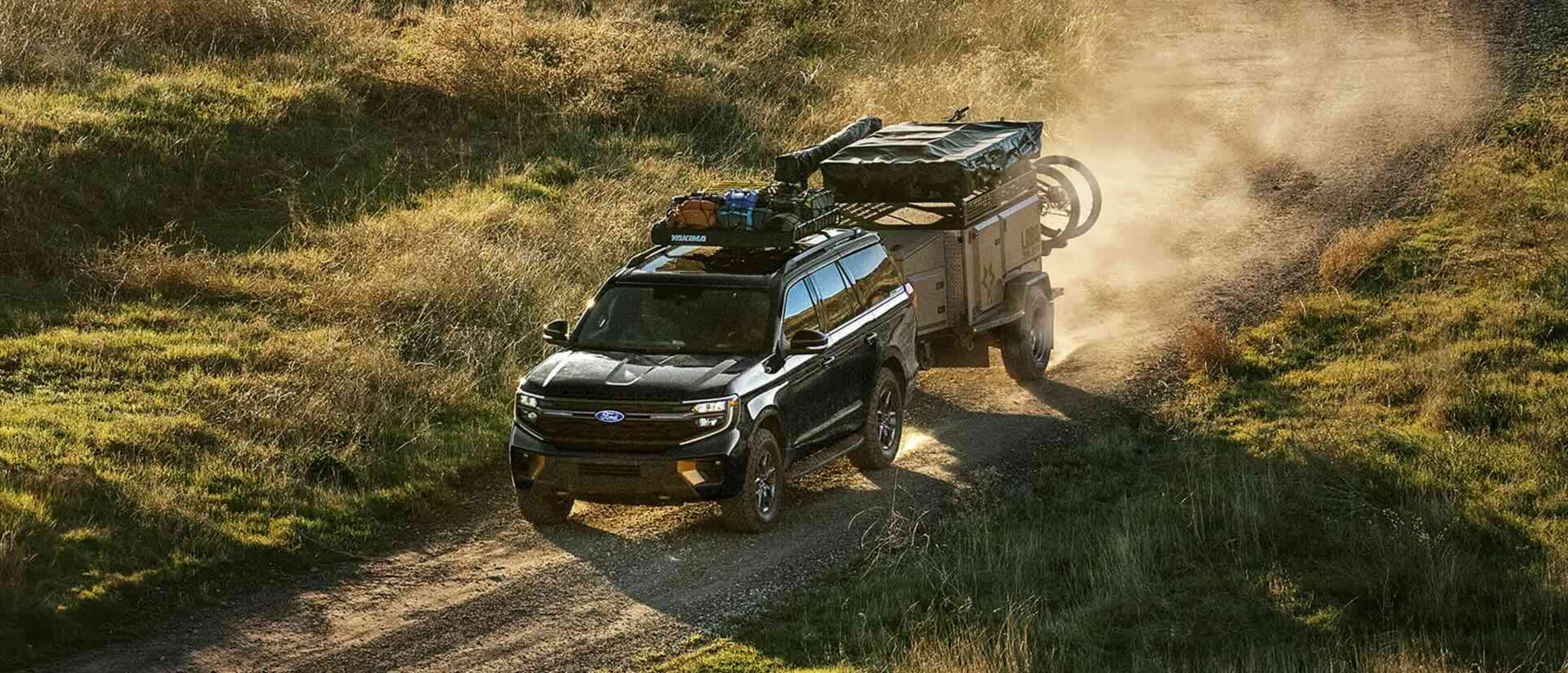 A black SUV towing a trailer along a dusty dirt road through a grassy, sunlit landscape.