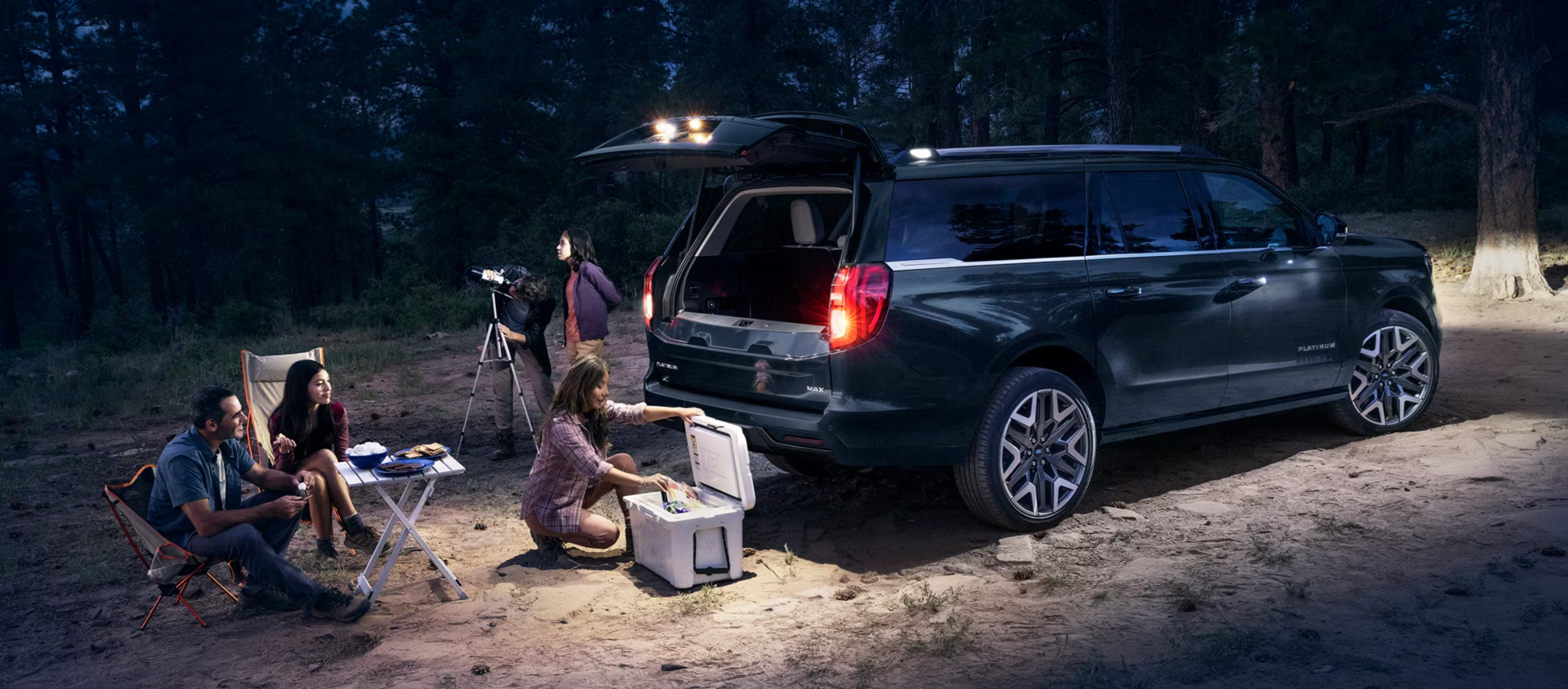 A group of people gathered around a cooler and a table next to an open SUV with a dark, wooded area illuminated by artificial light in the background.