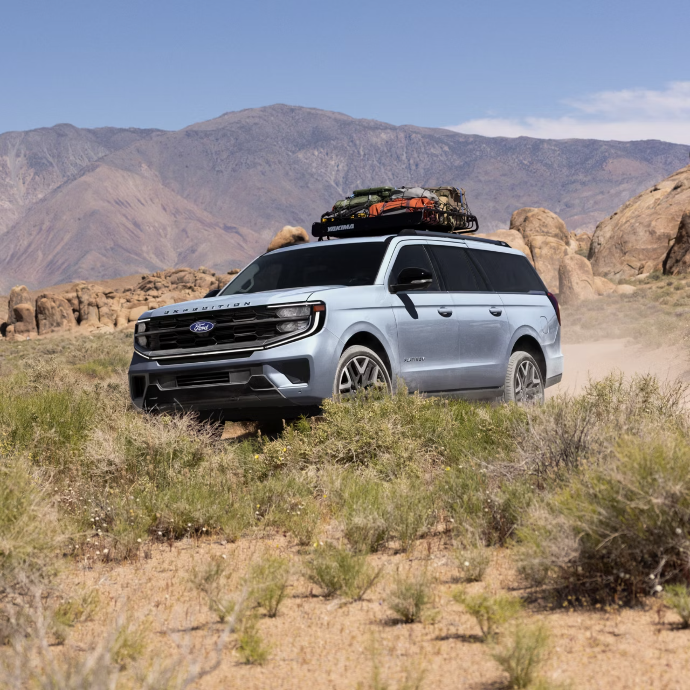 A light blue SUV with a roof rack loaded with gear drives on a dusty dirt road through a desert landscape with rocky hills and sparse vegetation under a clear blue sky.