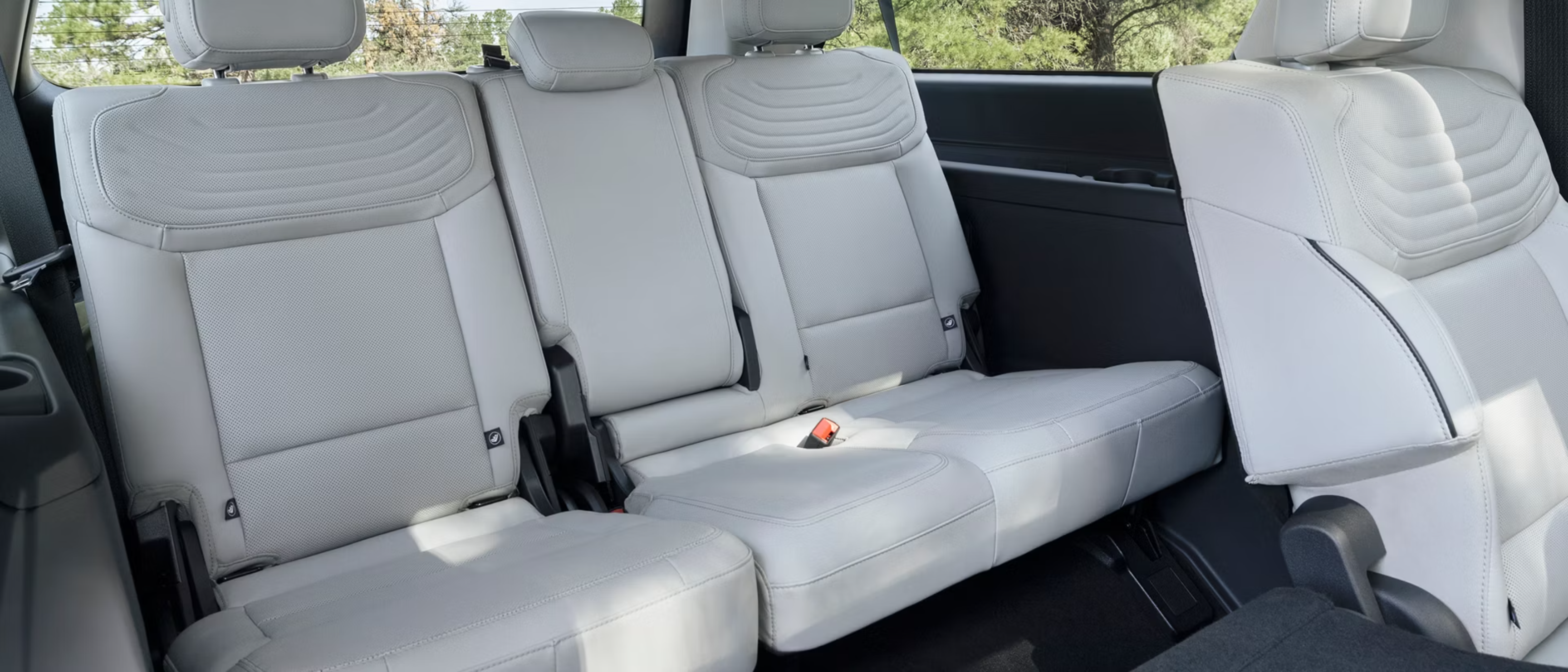 Interior view of a car's second and third rows with light gray leather seats, with a background of trees and sky visible through the windows.
