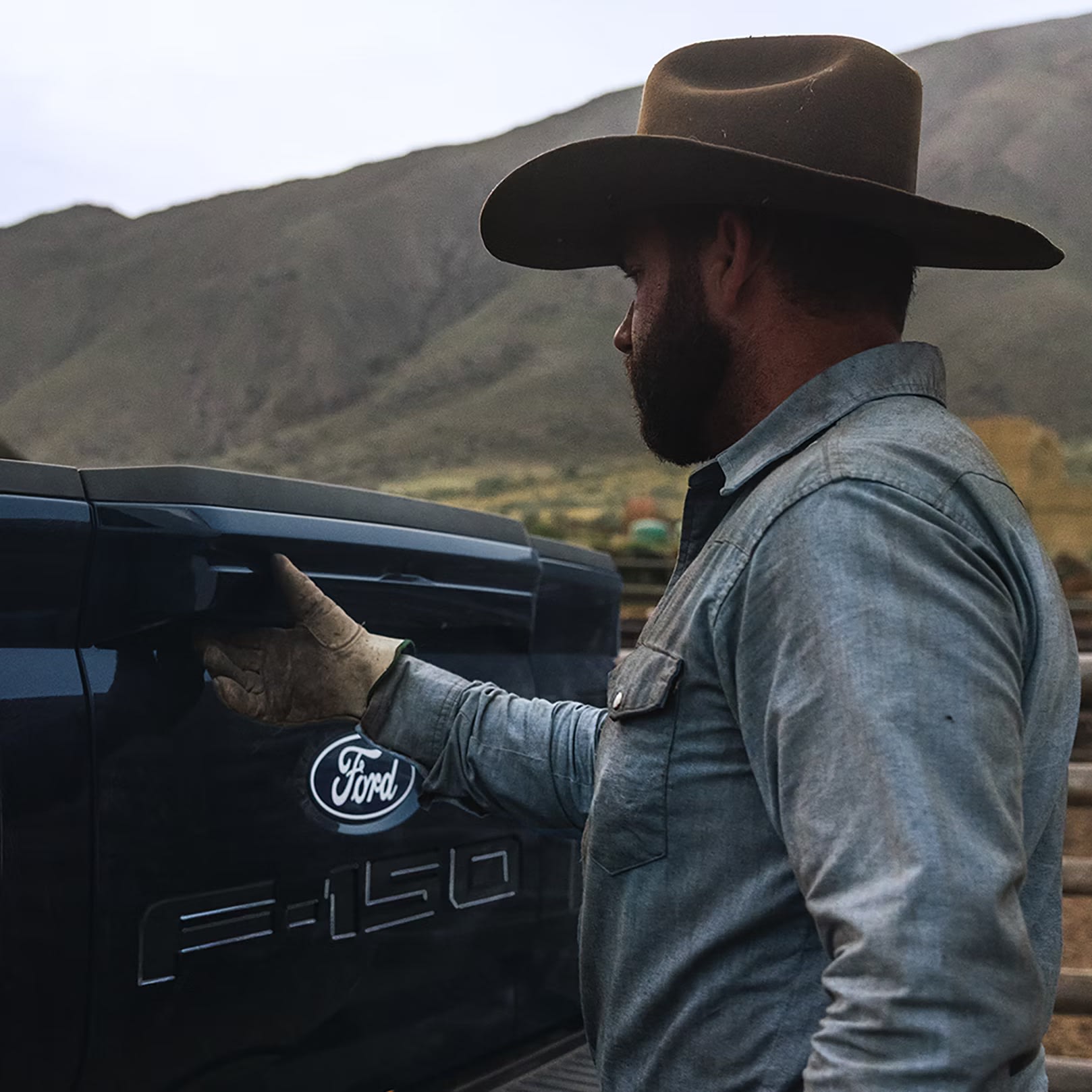 A man in a cowboy hat and work shirt opens the tailgate of a pickup truck with mountains in the background.