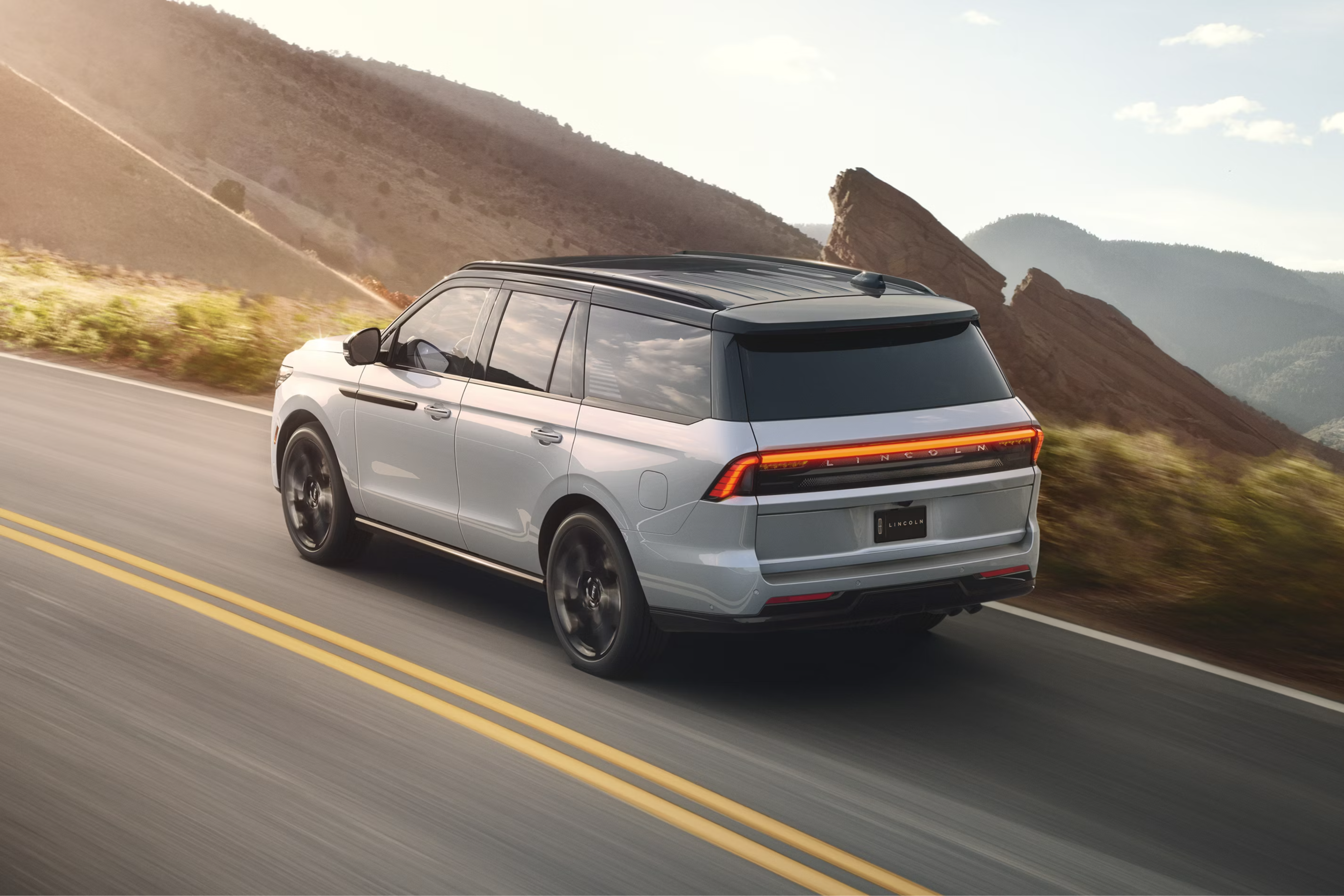 A white SUV drives along a winding mountain road under a bright, hazy sky.