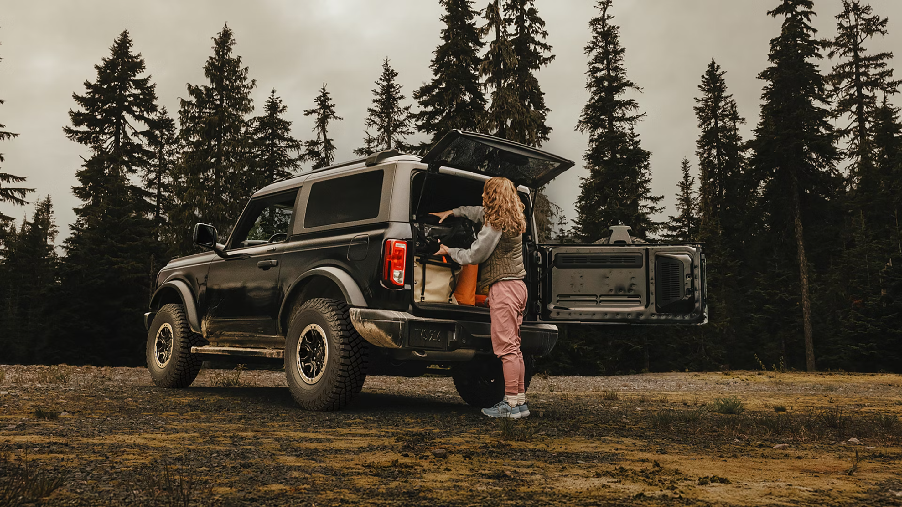 A woman loads gear into the back of a black SUV parked on a grassy clearing with tall evergreen trees in the background.