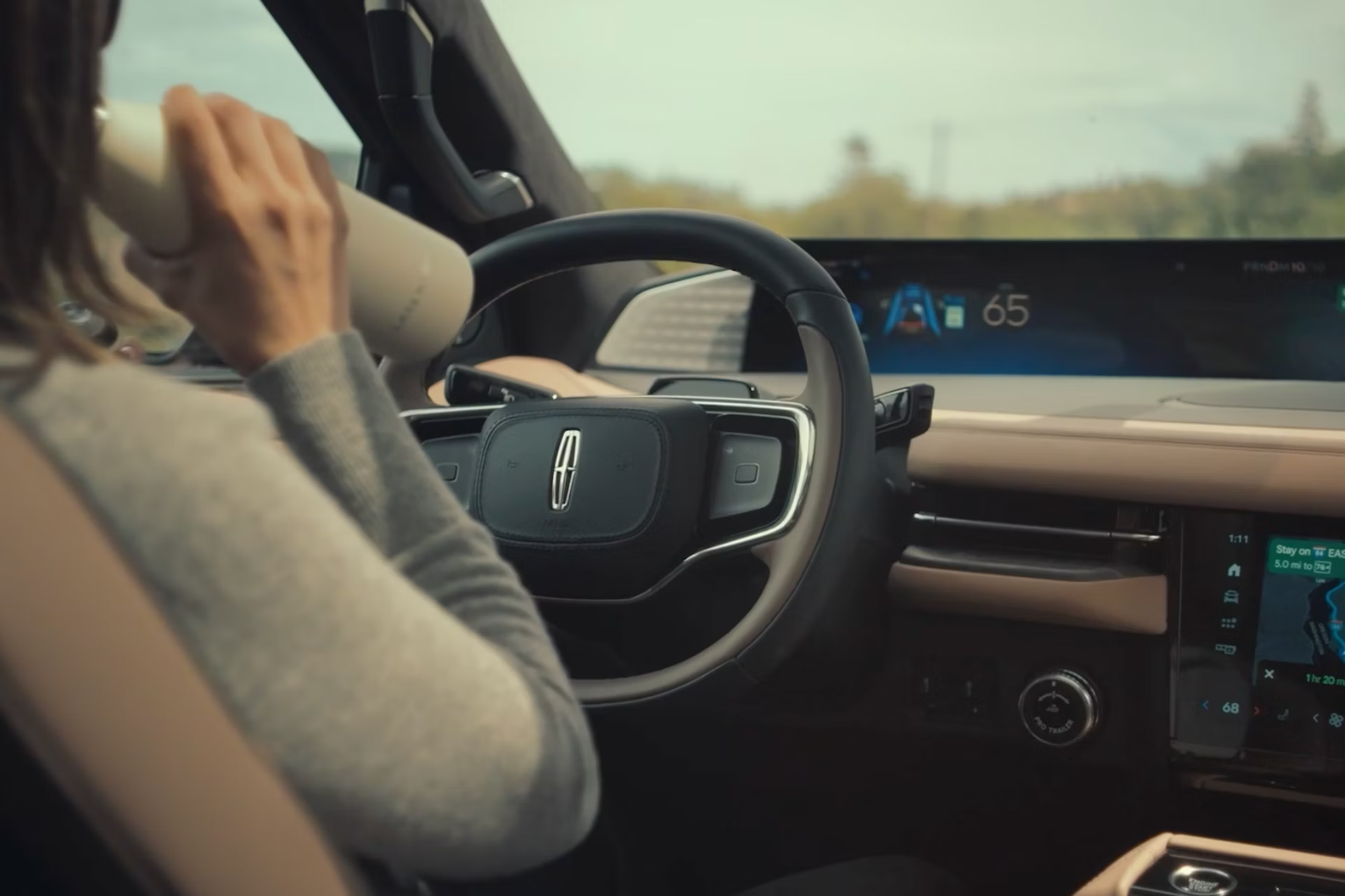 A person drinking from a bottle while driving a modern car with a digital dashboard showing speed and navigation, with a blurred landscape visible through the windshield.