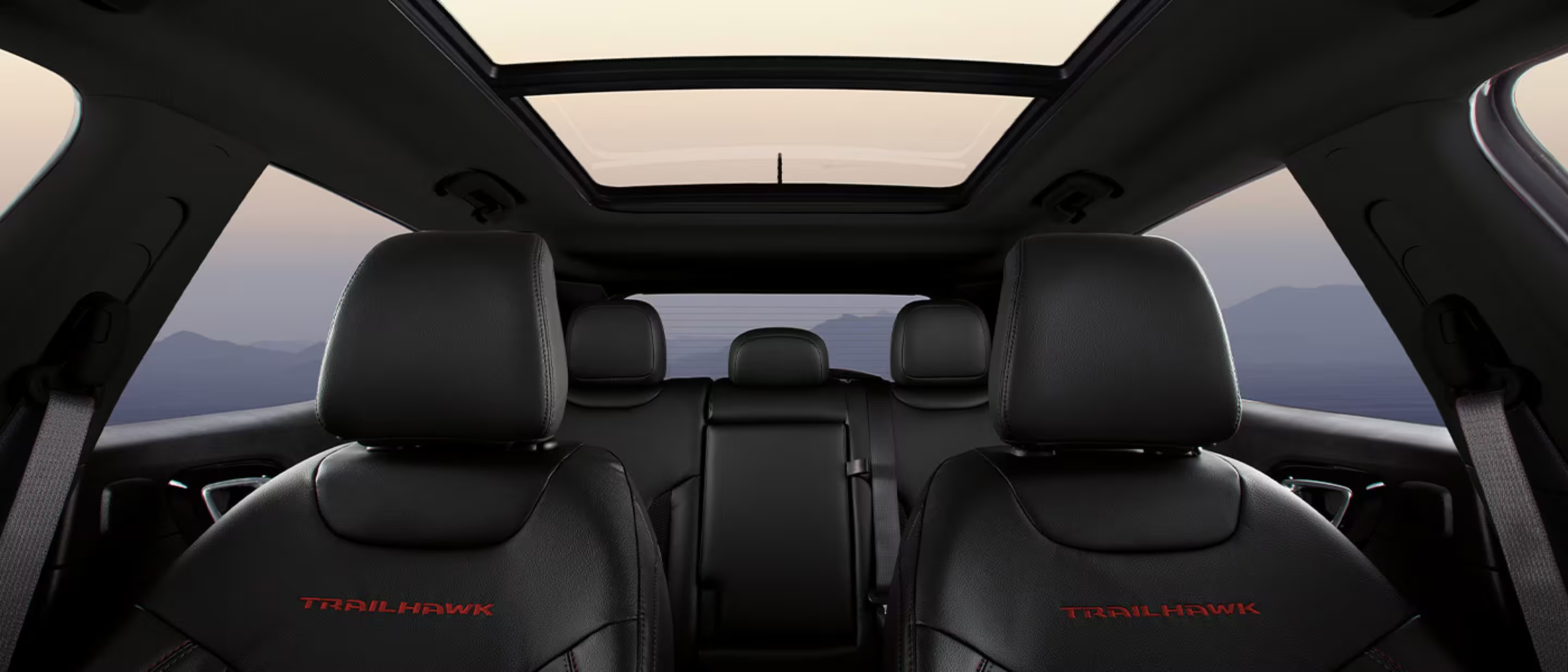Black leather car seats with red embroidery in foreground beneath a panoramic sunroof, with mountain landscape visible in background.