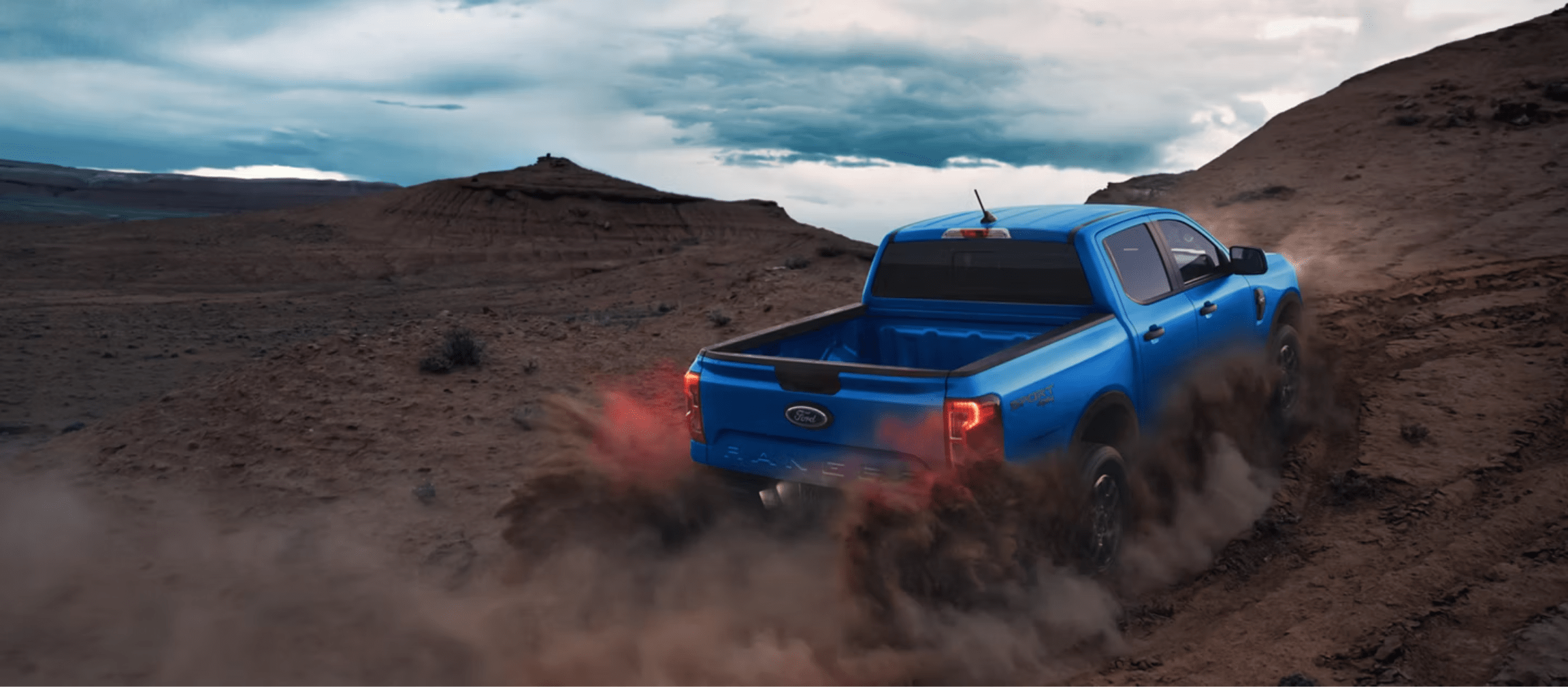 A blue pickup truck kicks up clouds of dust driving through a rugged desert landscape under a cloudy sky.
