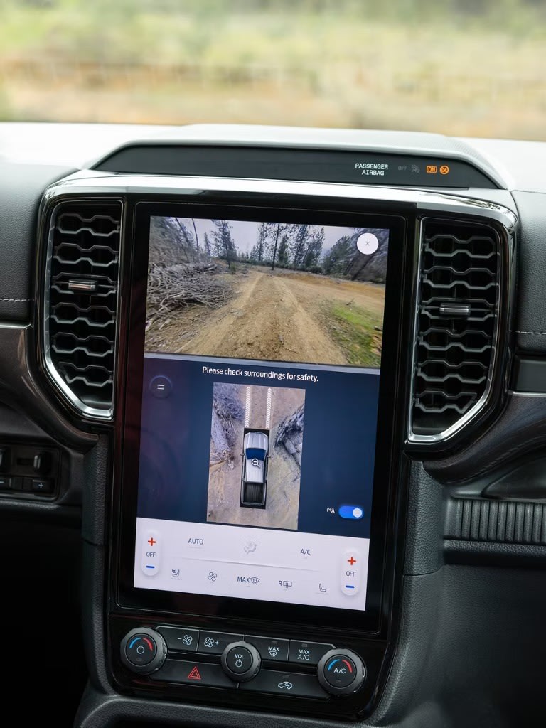 A large touchscreen displays a rearview camera feed of a dirt road with trees in the background, while the foreground shows the vehicle's dashboard.