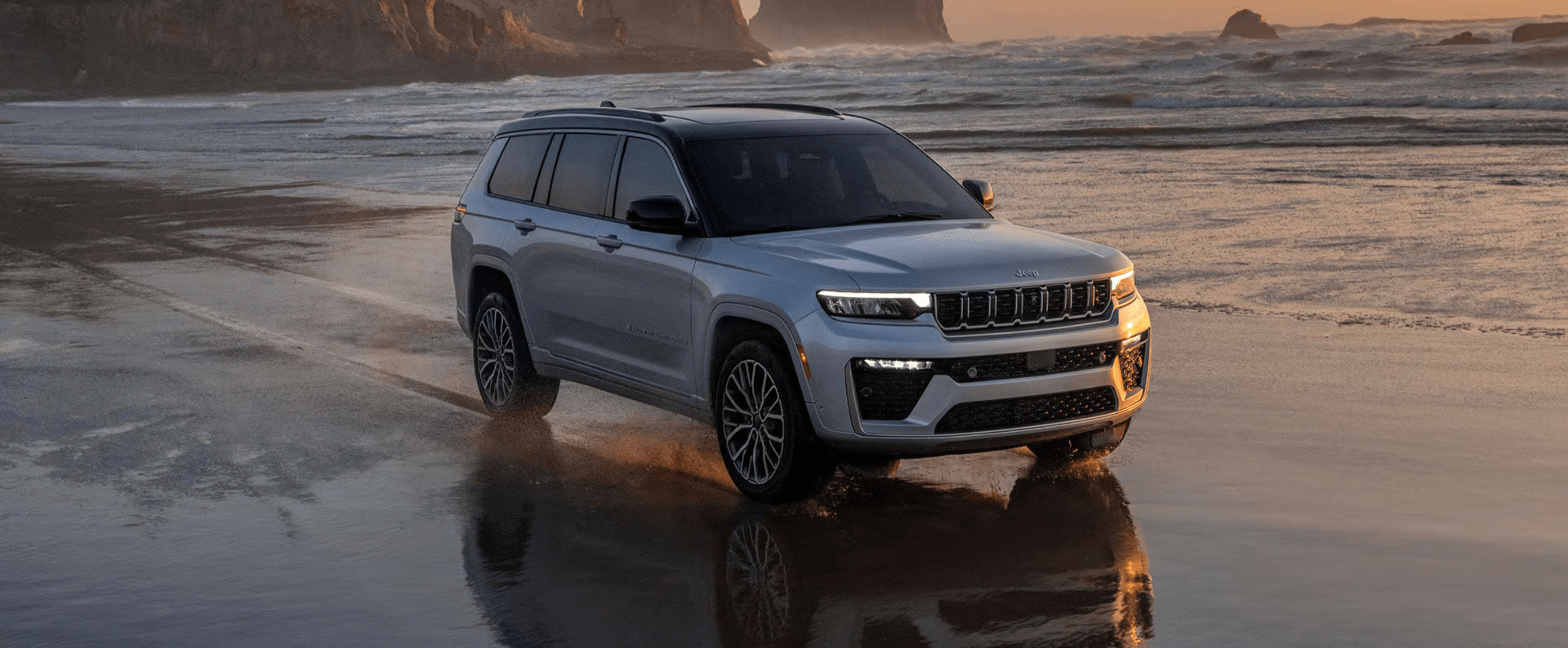 Silver SUV on wet sandy beach with ocean waves and rocky cliffs in background.