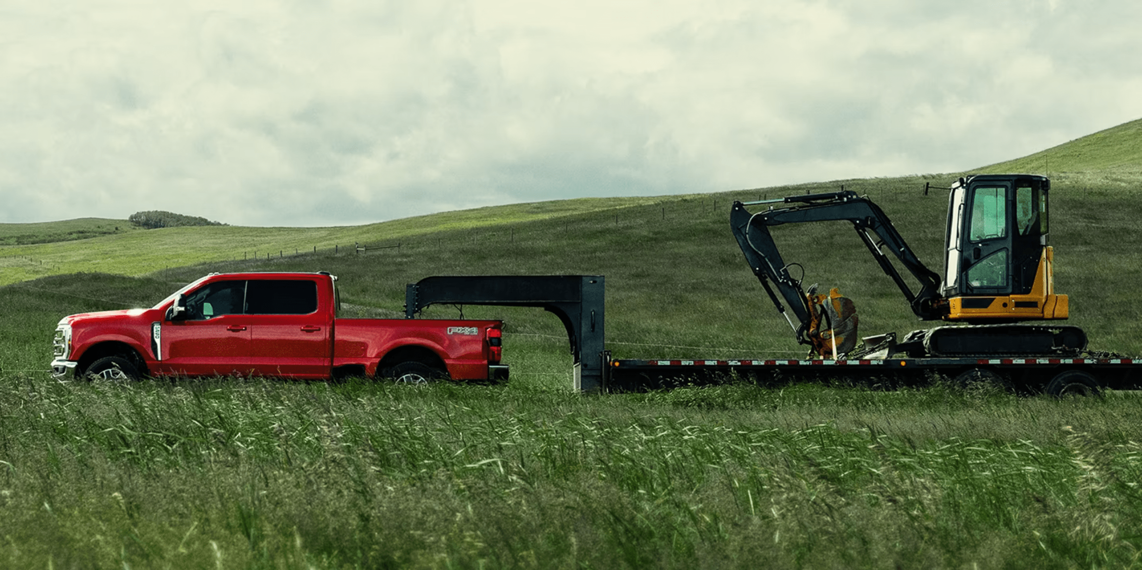 Red pickup truck towing yellow excavator on flatbed trailer in grassy field with green hills and cloudy sky.