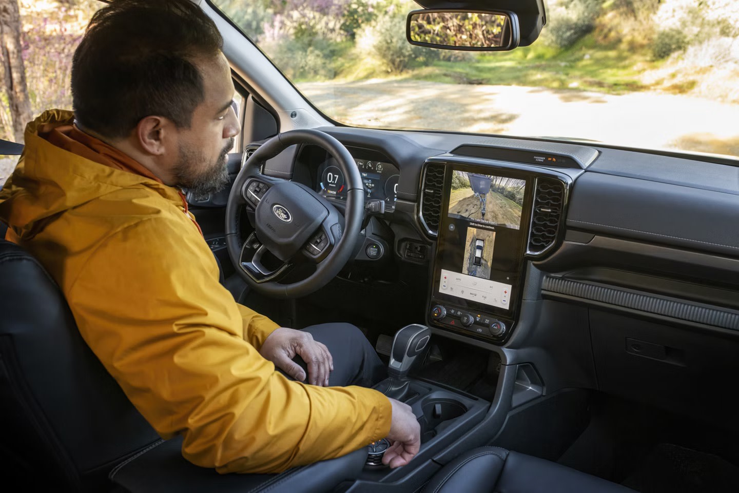 A person in a yellow jacket sits in the driver's seat of a modern vehicle, looking at the large touchscreen display which shows a view of the road ahead and a camera feed, while the background reveals a dirt road surrounded by trees.