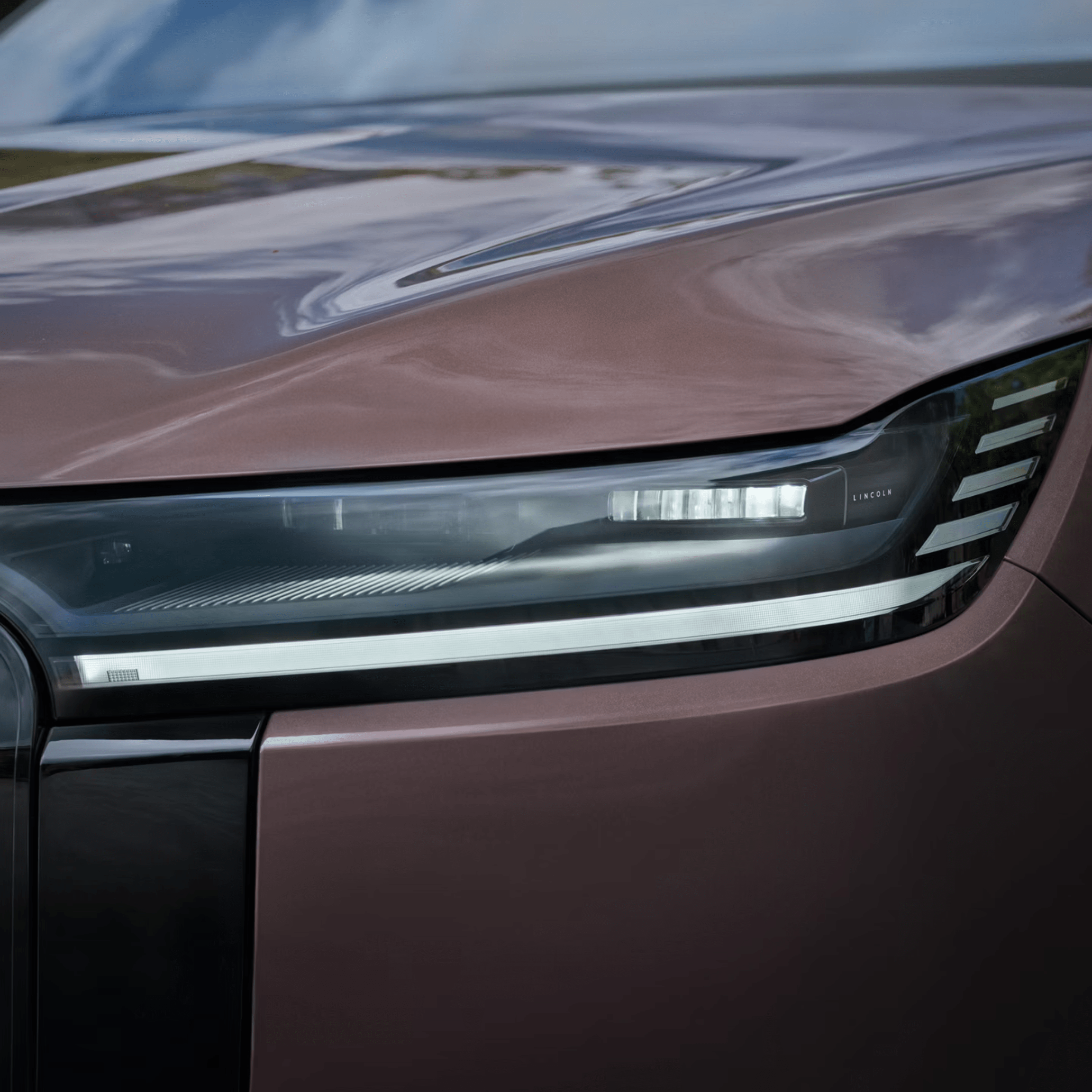 Close-up of a brown car's headlight and hood, with blurred sky reflection.