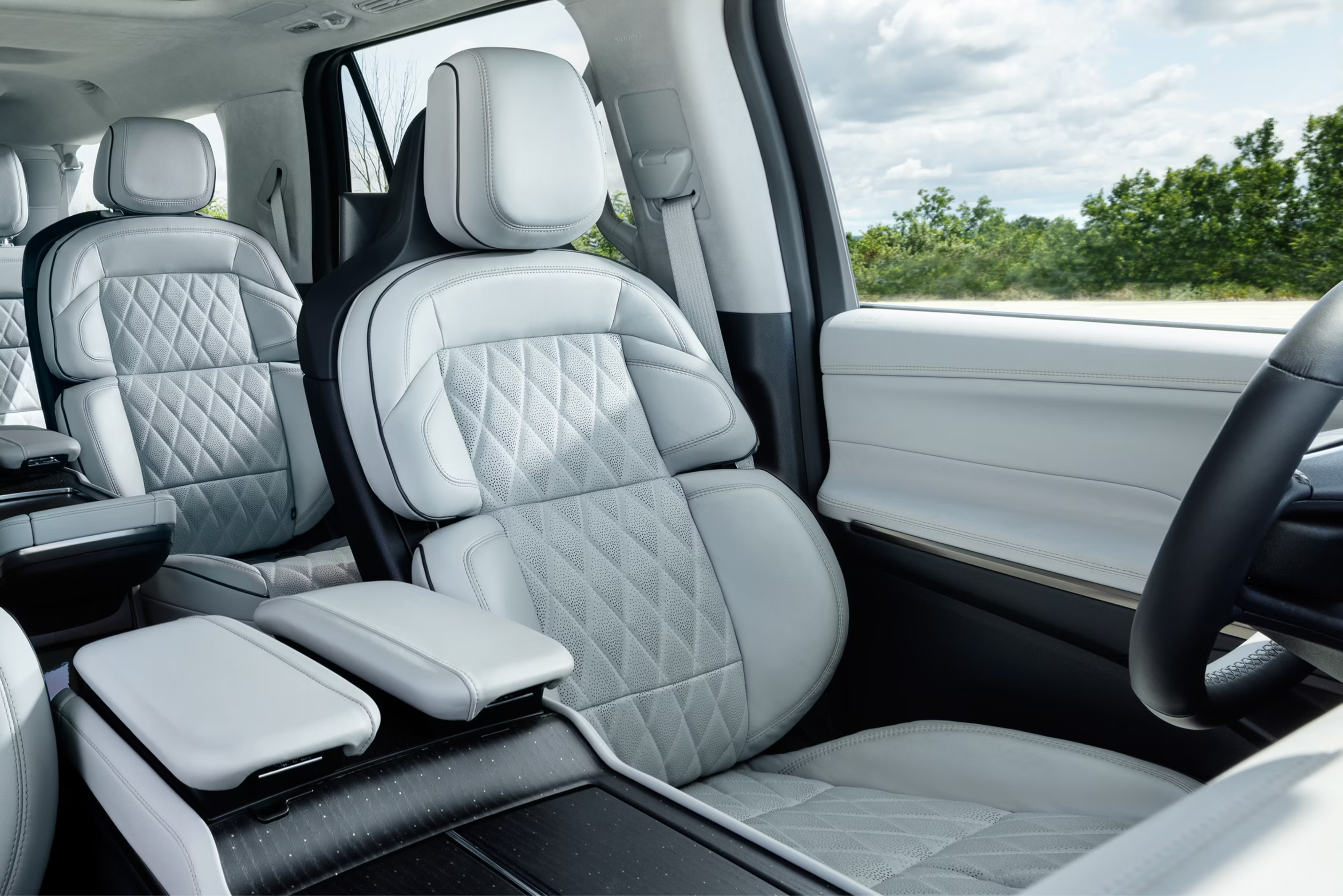 White quilted leather car seats fill the foreground, with green trees and a cloudy sky visible through the window in the background.