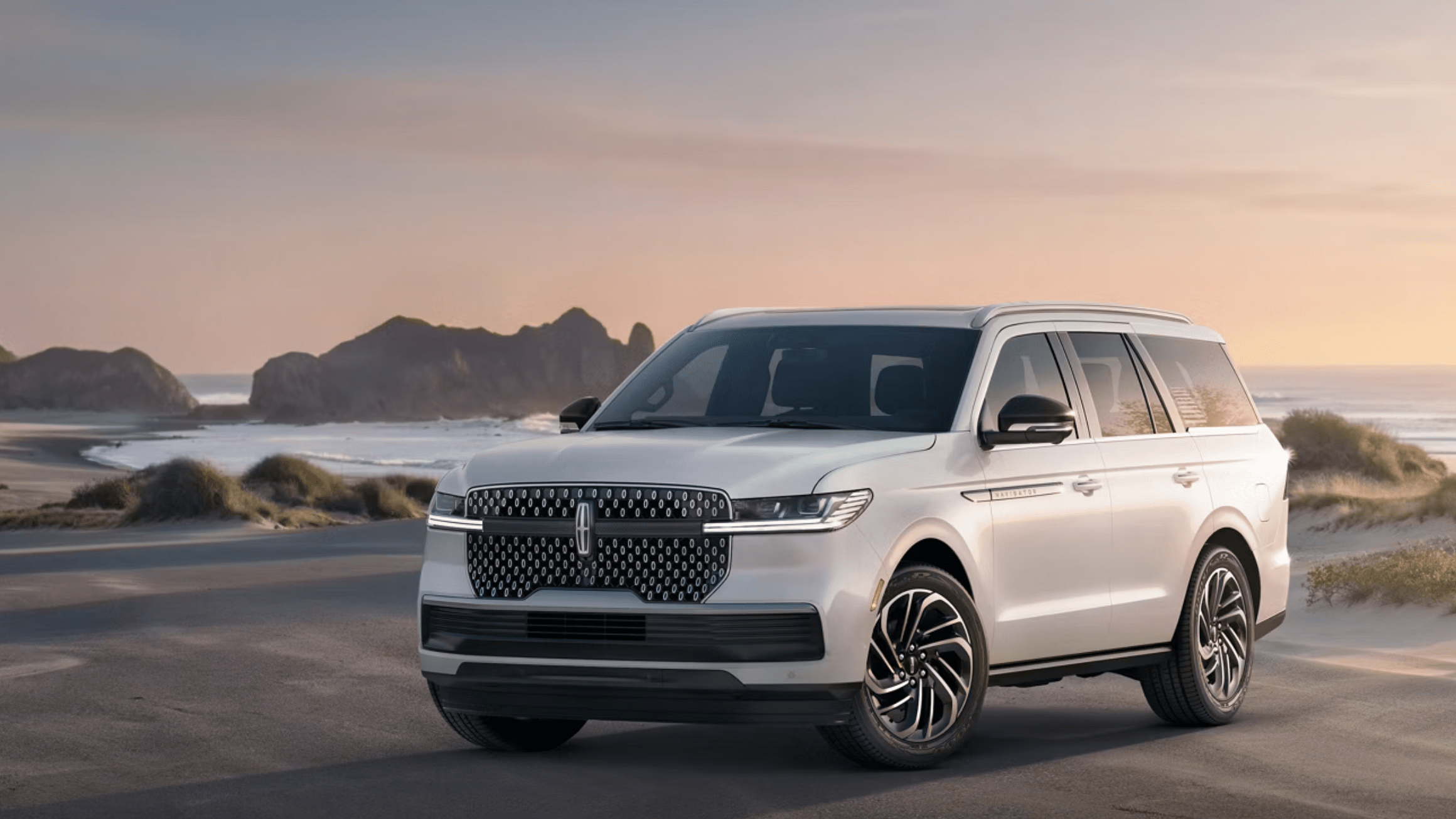 A white SUV is parked on a sandy beach with rocky outcrops and the ocean in the background under a soft, hazy sky.