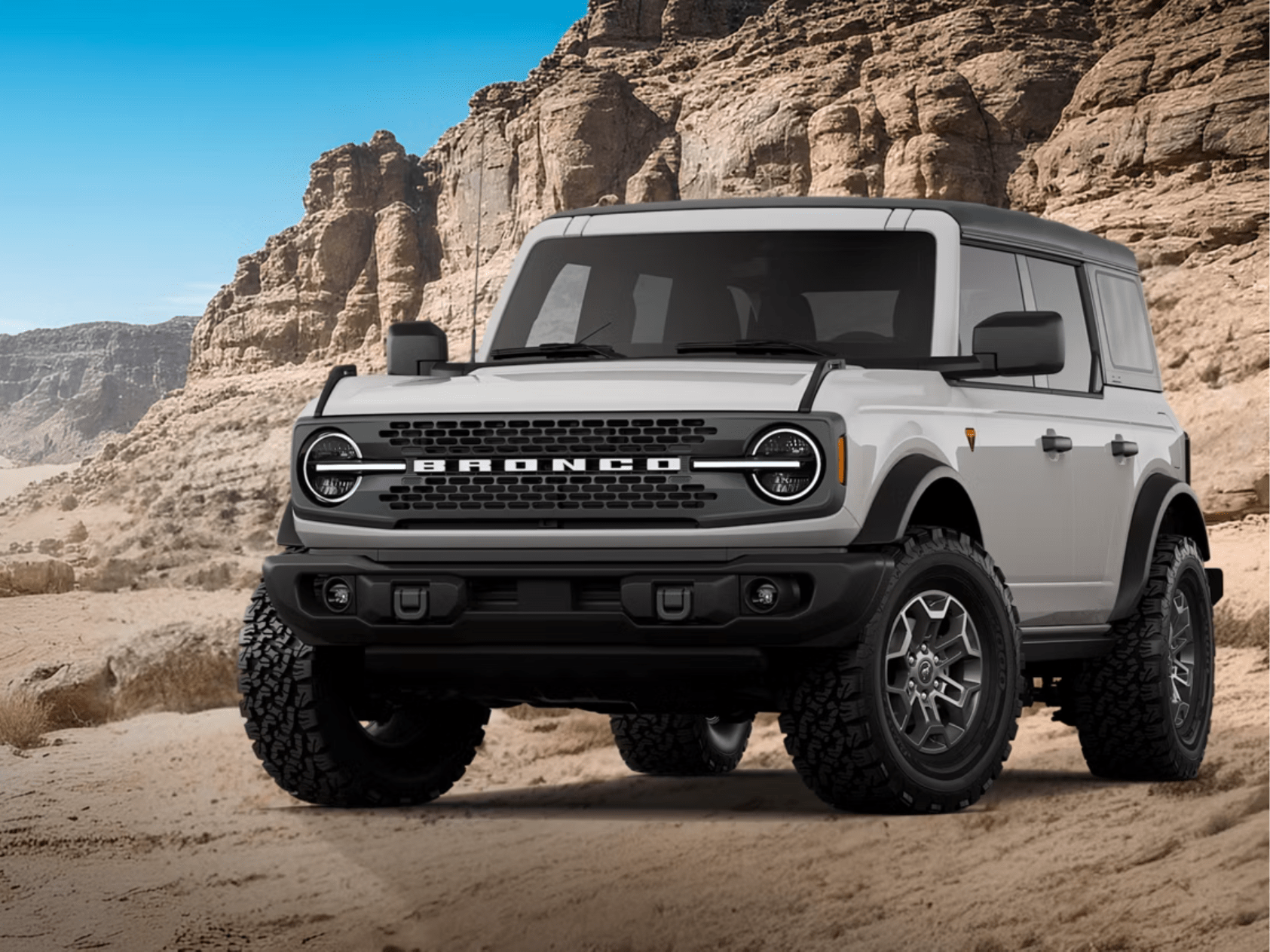 A white off-road SUV sits on a sandy desert floor against a backdrop of towering sandstone cliffs and a clear blue sky.