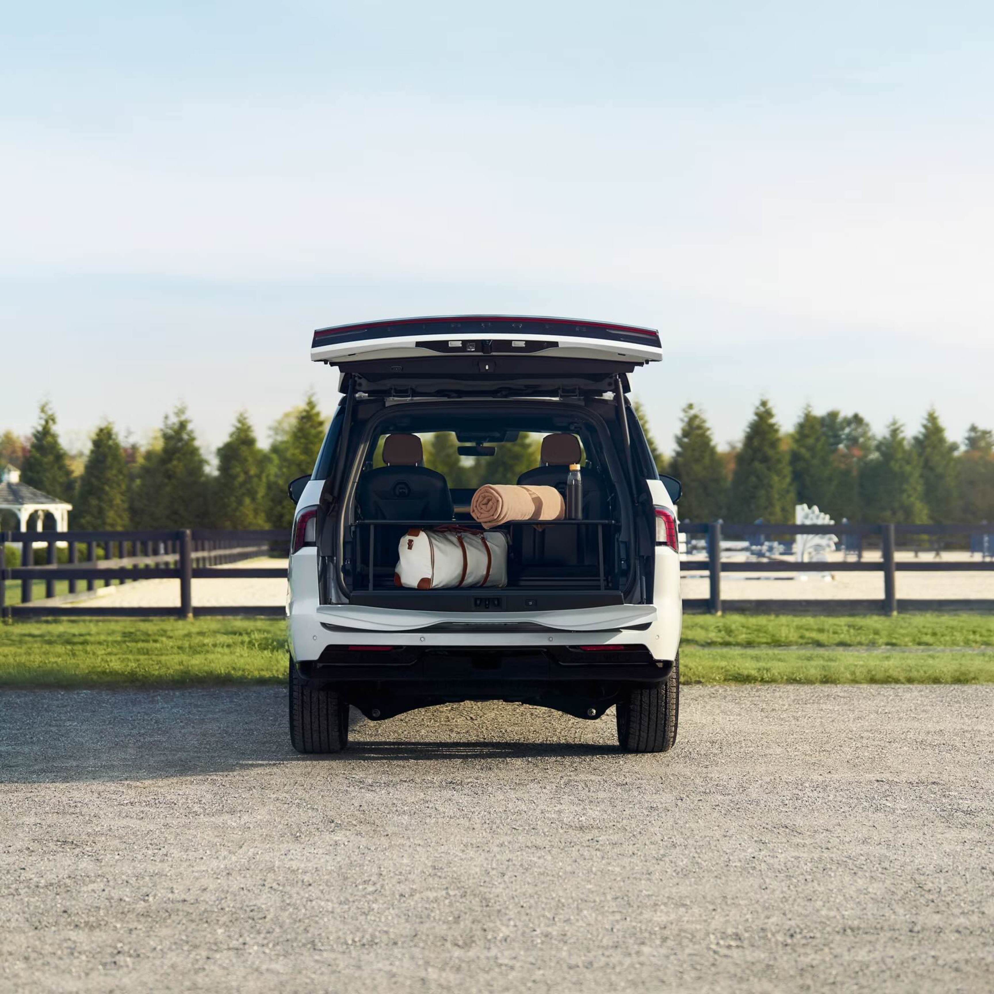 White SUV with open trunk on gravel driveway with trees and field in background.