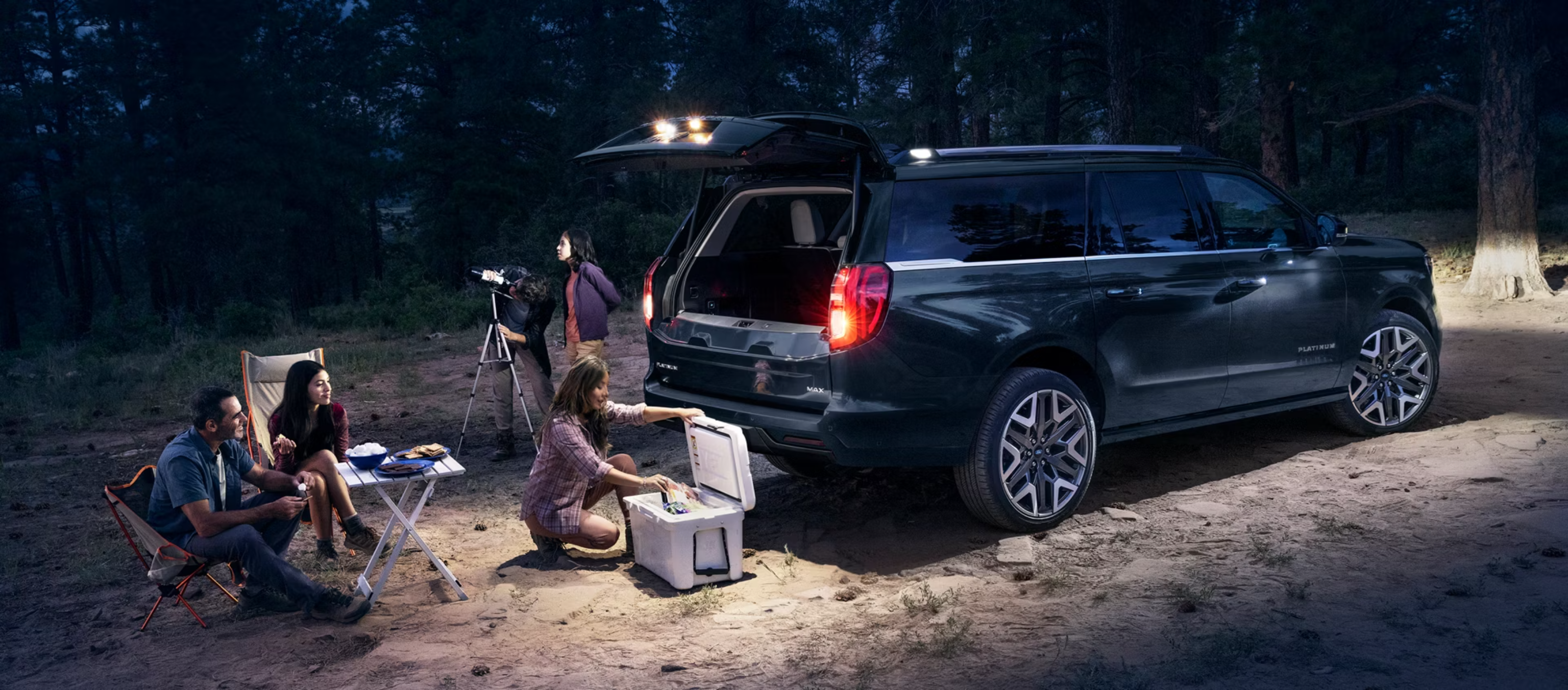 People enjoying a campsite next to a dark SUV with an open trunk in a wooded area at night.