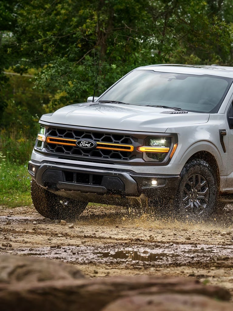 A silver truck splashes through a muddy trail with lush green trees in the background.