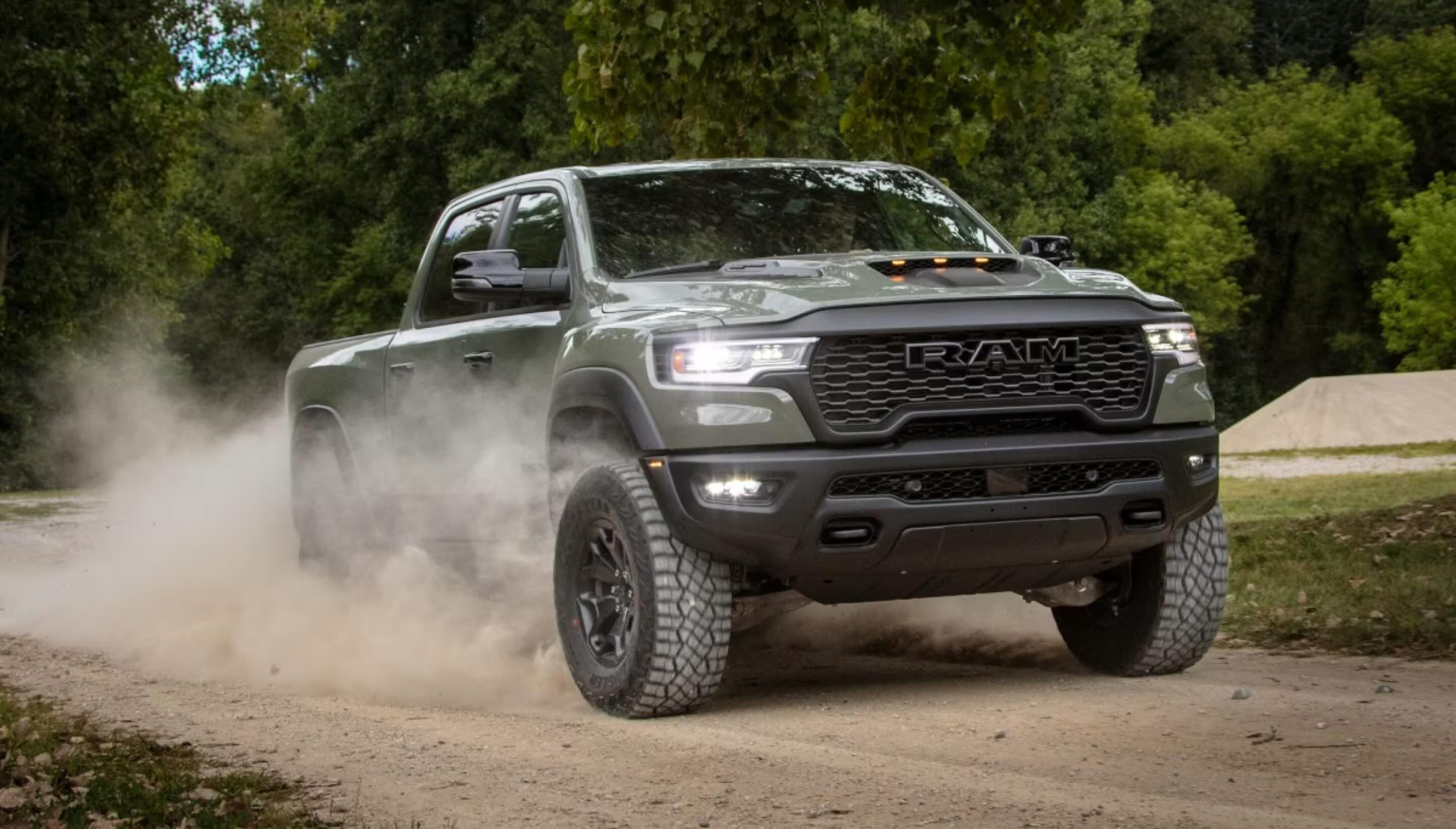 A green truck kicks up dust as it drives on a dirt road with a wooded background.