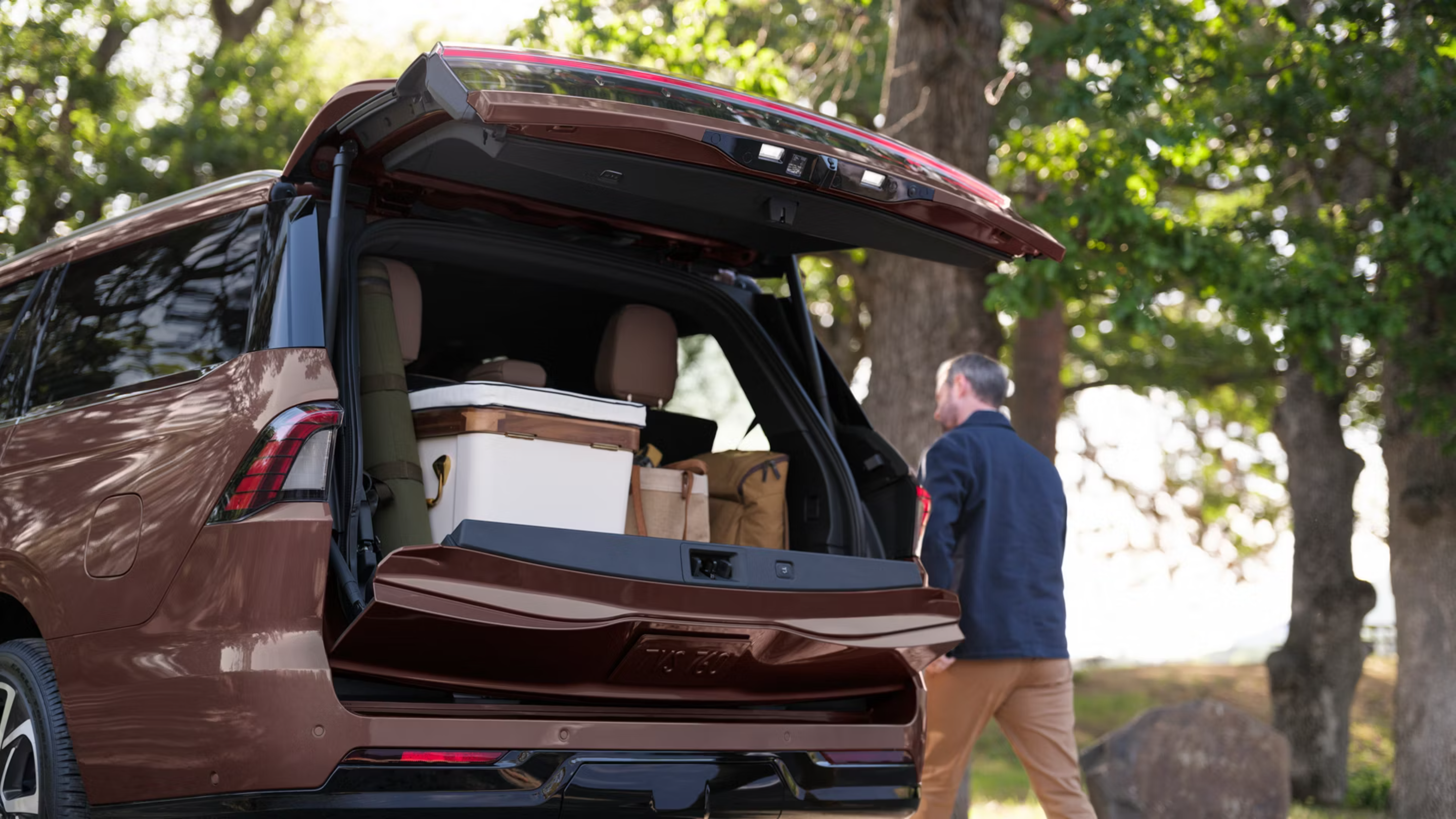 A brown SUV with its trunk open and filled with gear sits in the foreground as a man walks away toward a backdrop of lush green trees.