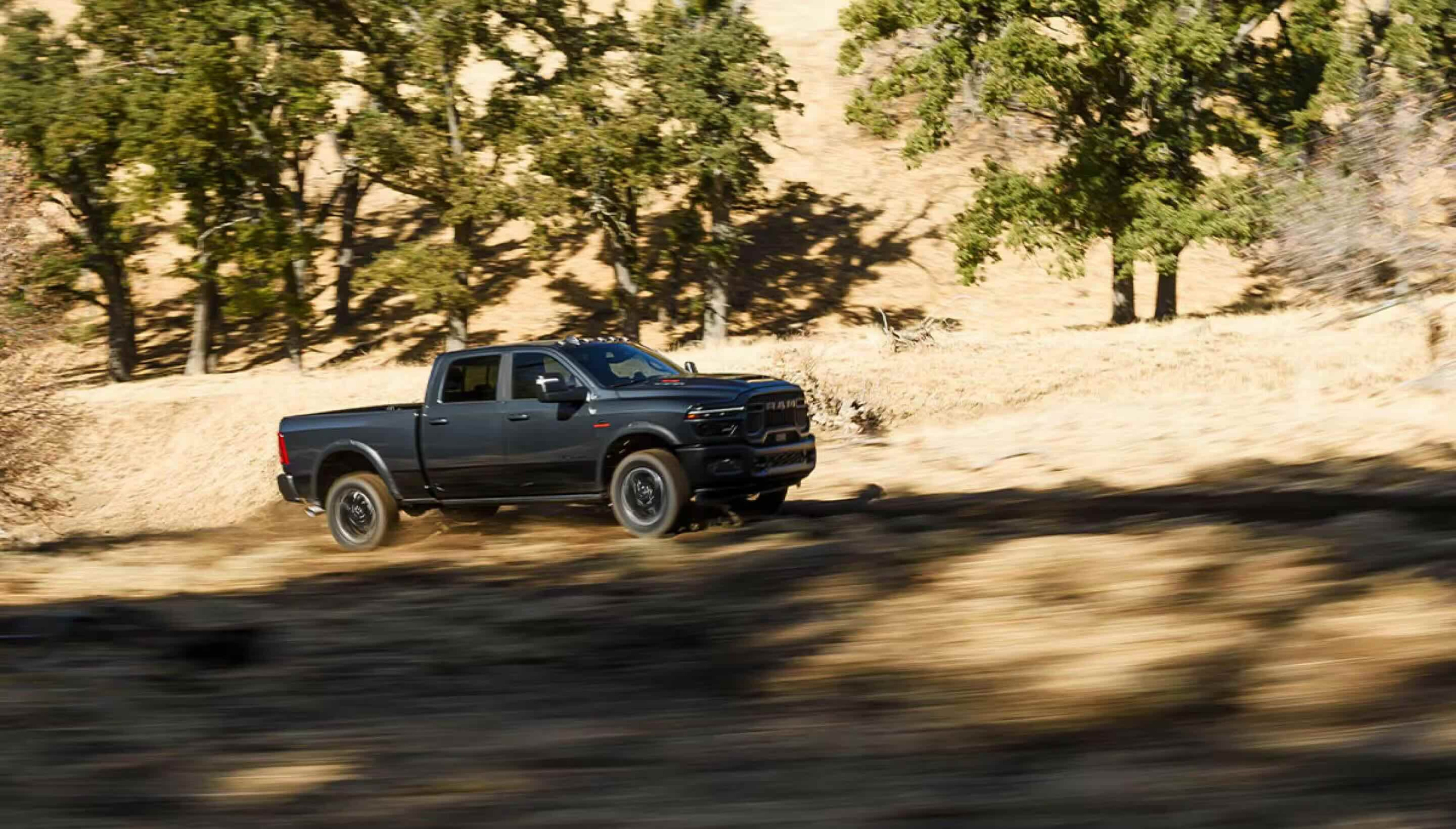 A dark gray pickup truck drives on a dirt road in the foreground, with a dry, wooded hillside in the background.