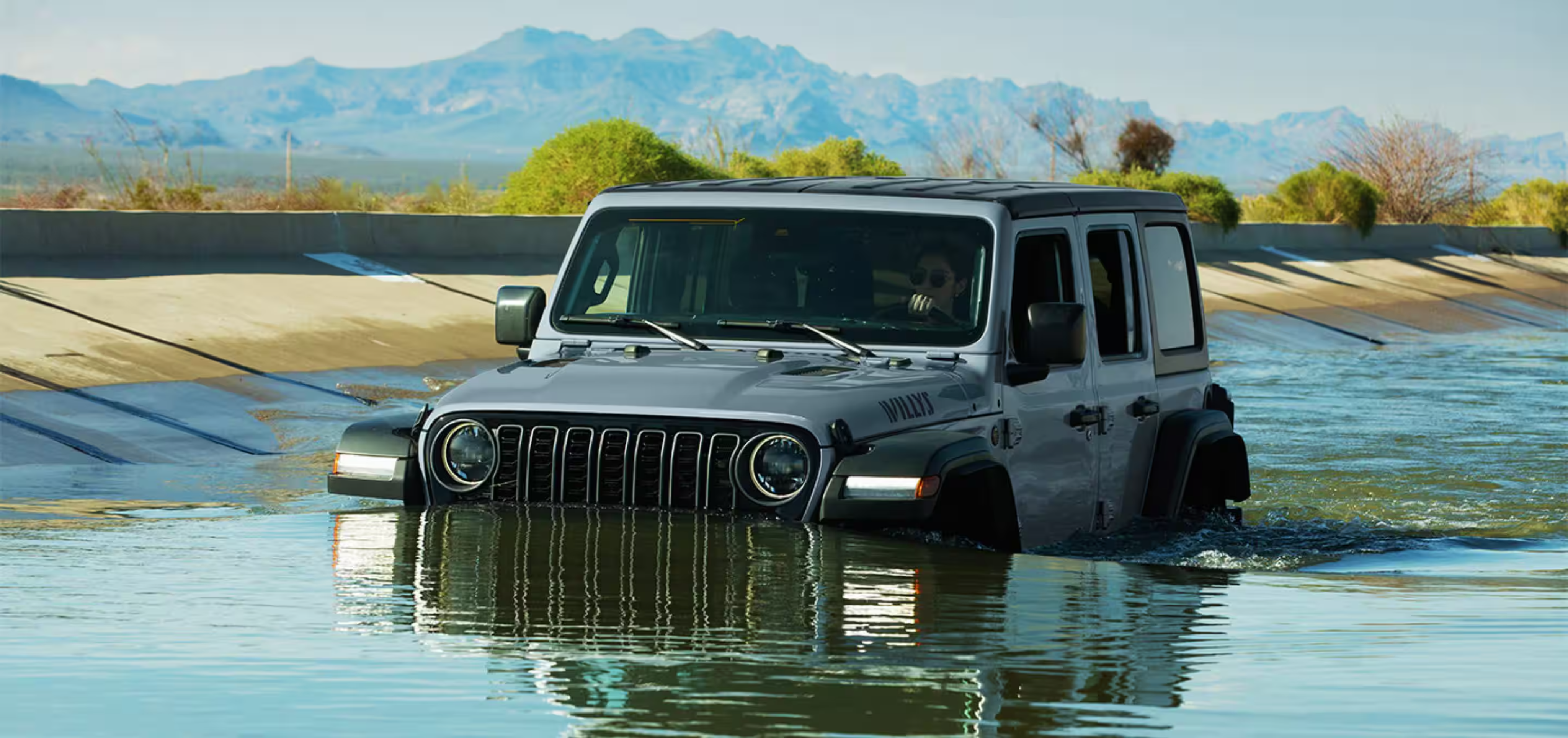 A grey SUV driving through shallow water in a concrete channel with mountains and greenery in the background.