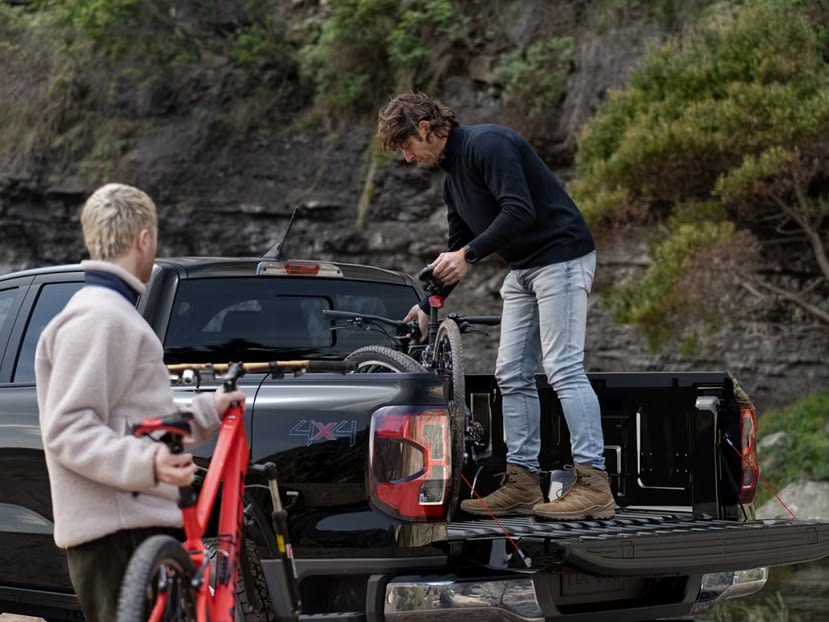 Two men loading a bicycle into the bed of a pickup truck with a rocky, wooded hillside in the background.
