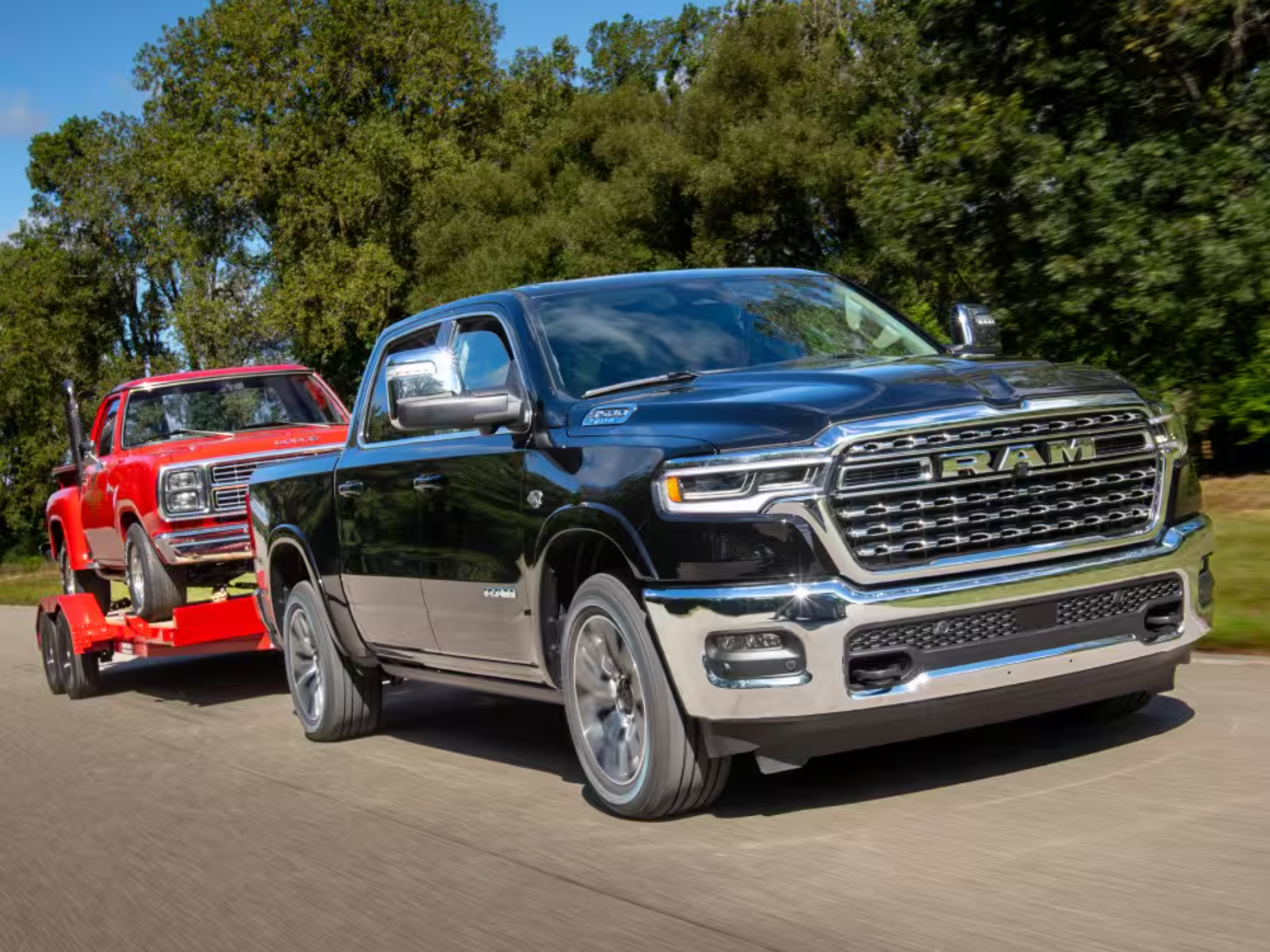 A black truck towing a red vintage truck on a trailer down a road with trees in the background.