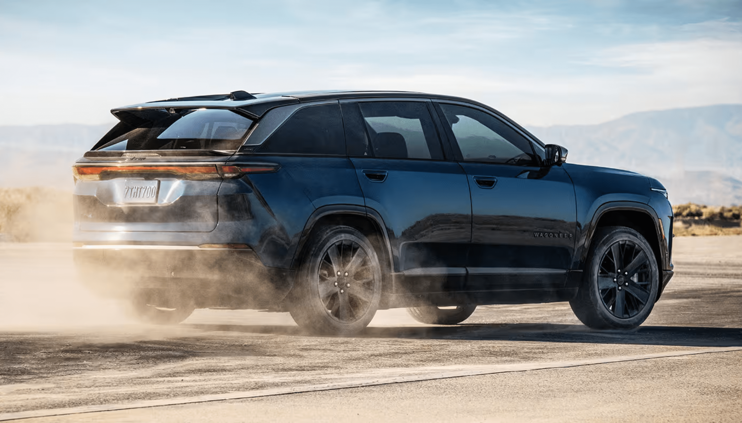 A dark blue SUV driving on a paved road with a desert landscape and mountains in the background.