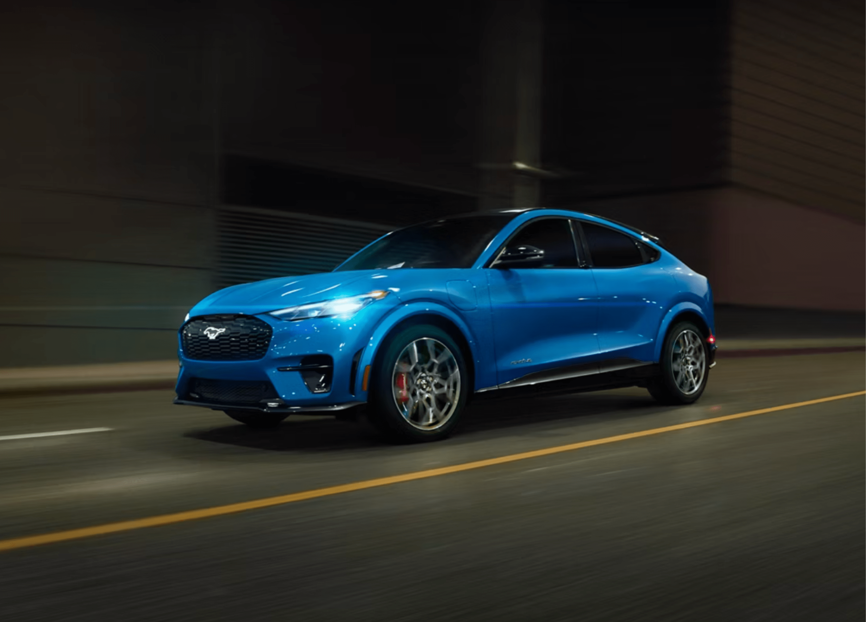 A bright blue electric SUV drives down a dark, wet road with blurred lights in the background.