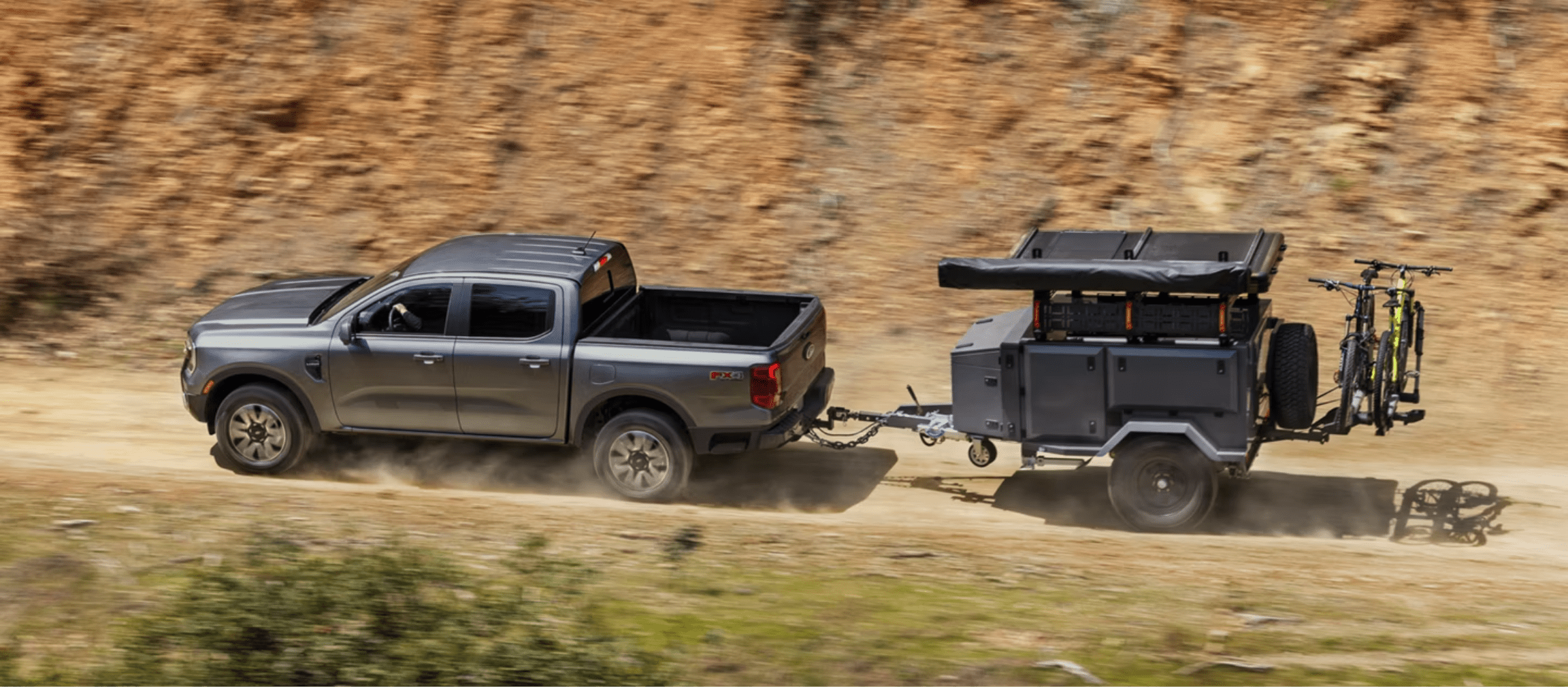 A grey pickup truck pulling a camping trailer with bicycles attached on a dusty dirt road with a blurred rocky hillside in the background.
