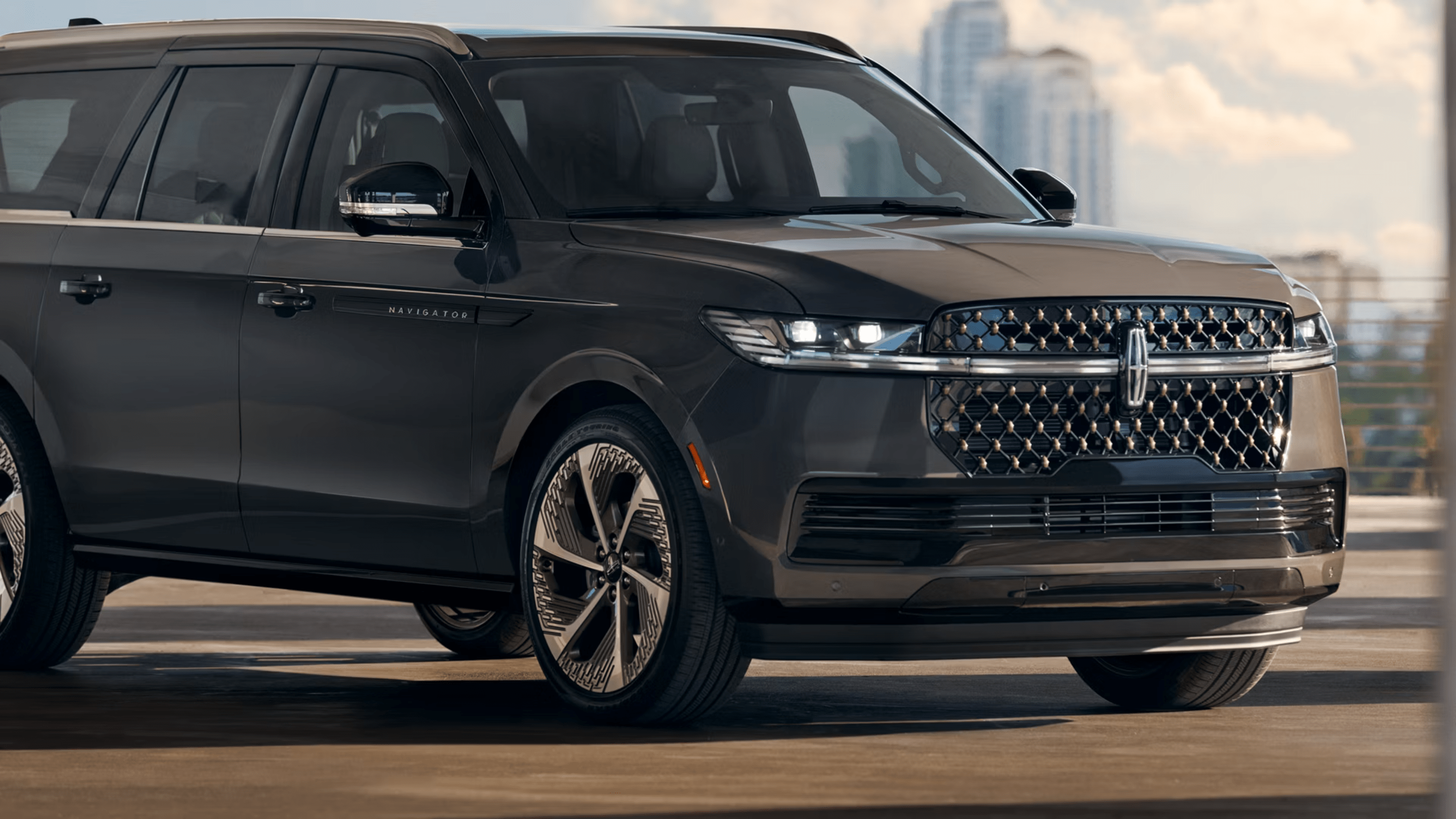 A large dark gray luxury SUV parked on a paved surface with blurred city buildings in the background.