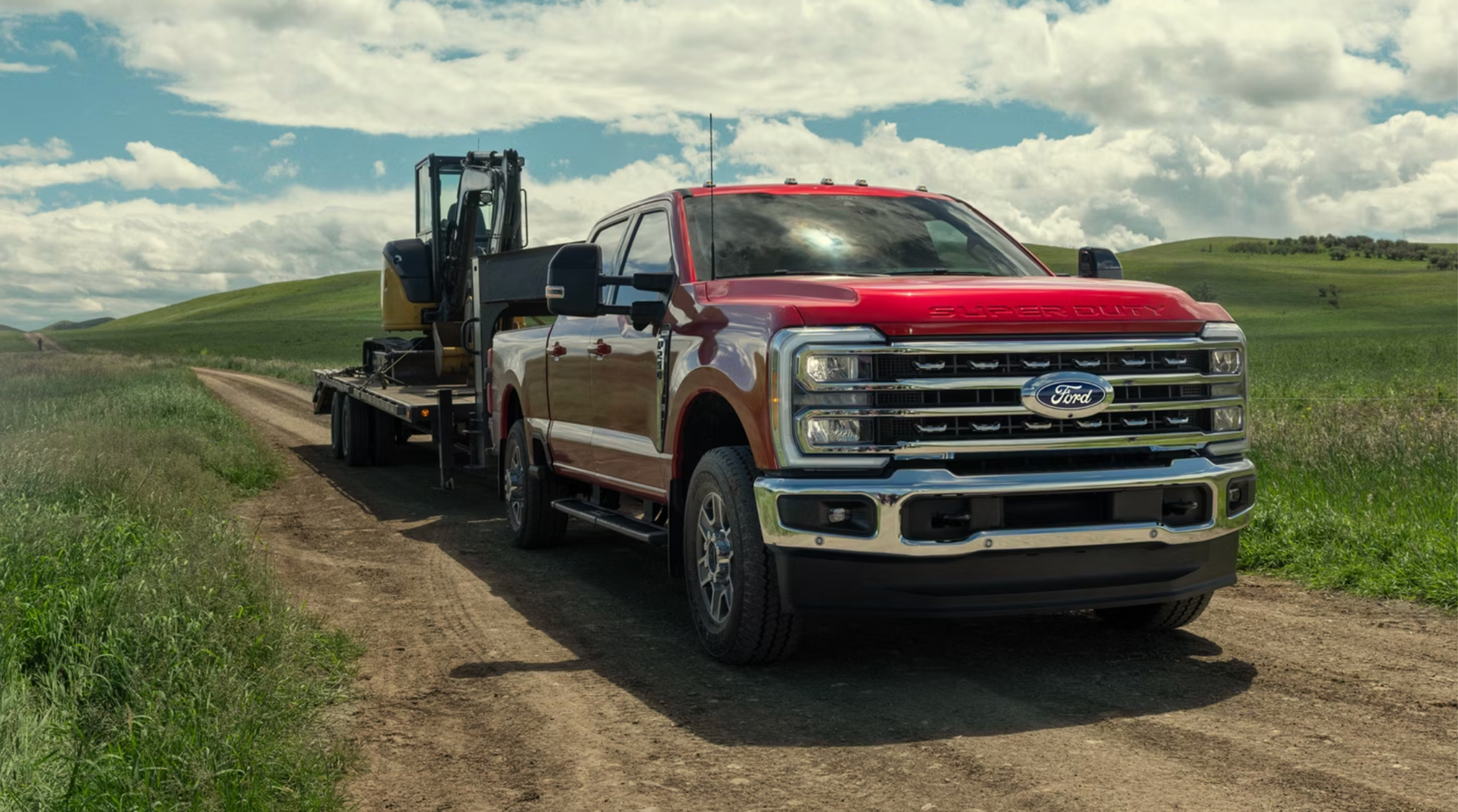 Red truck towing heavy machinery on dirt road surrounded by green hills under cloudy sky.