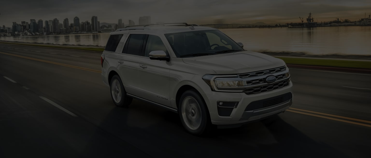 a white ford expedition vehicle driving down a road near a cityscape with a lake and a cityscape in the background