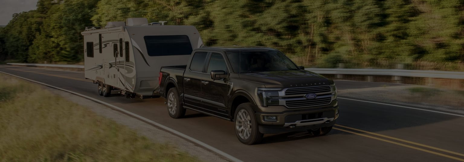 Image: A large pickup truck towing a recreational vehicle trailer on a winding road surrounded by lush, forested scenery.