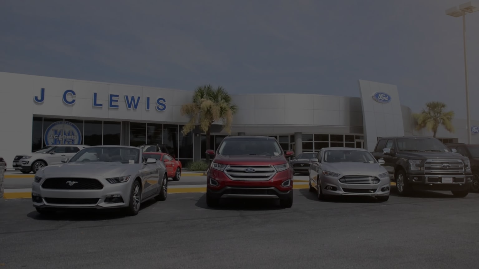 a group of cars parked in front of a j c lewis dealership with palm trees in the background