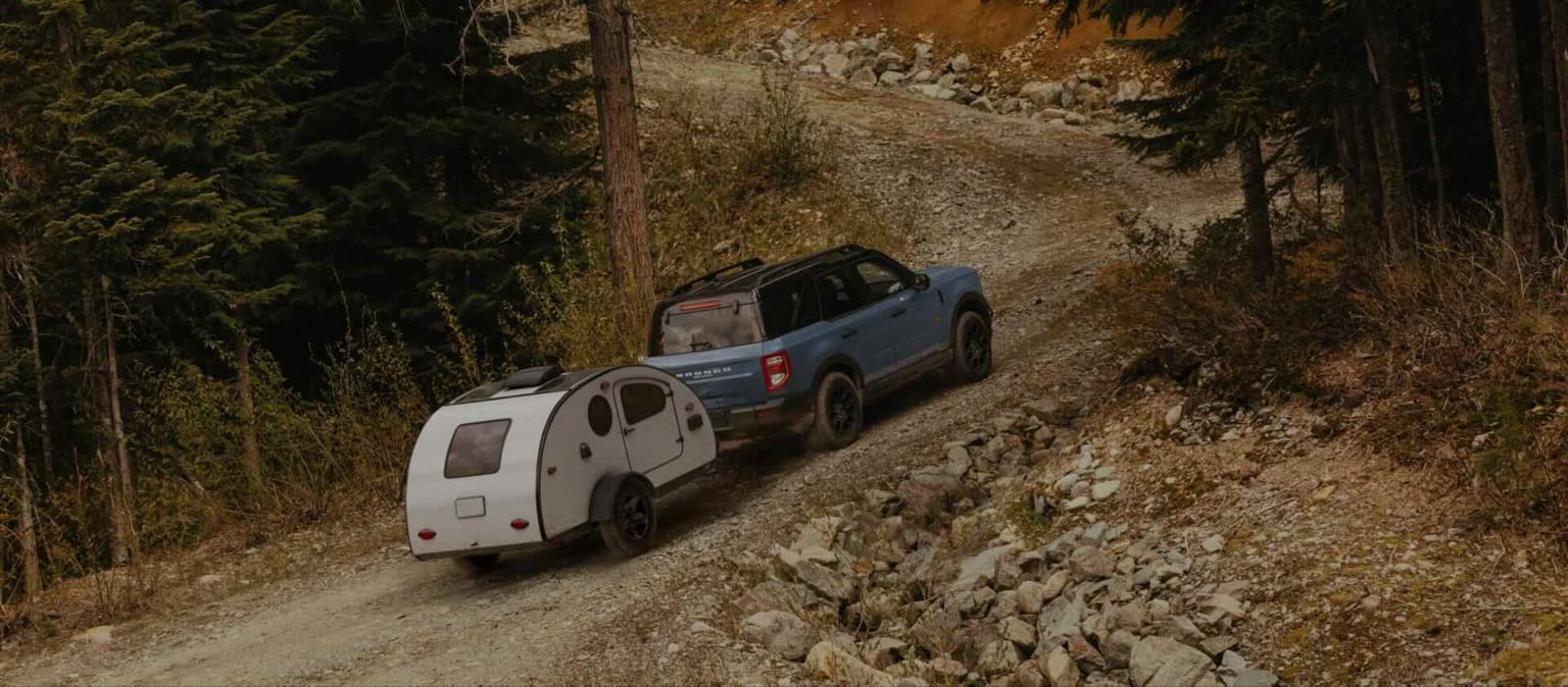 A blue SUV towing a white teardrop trailer up a rocky, wooded mountain road.