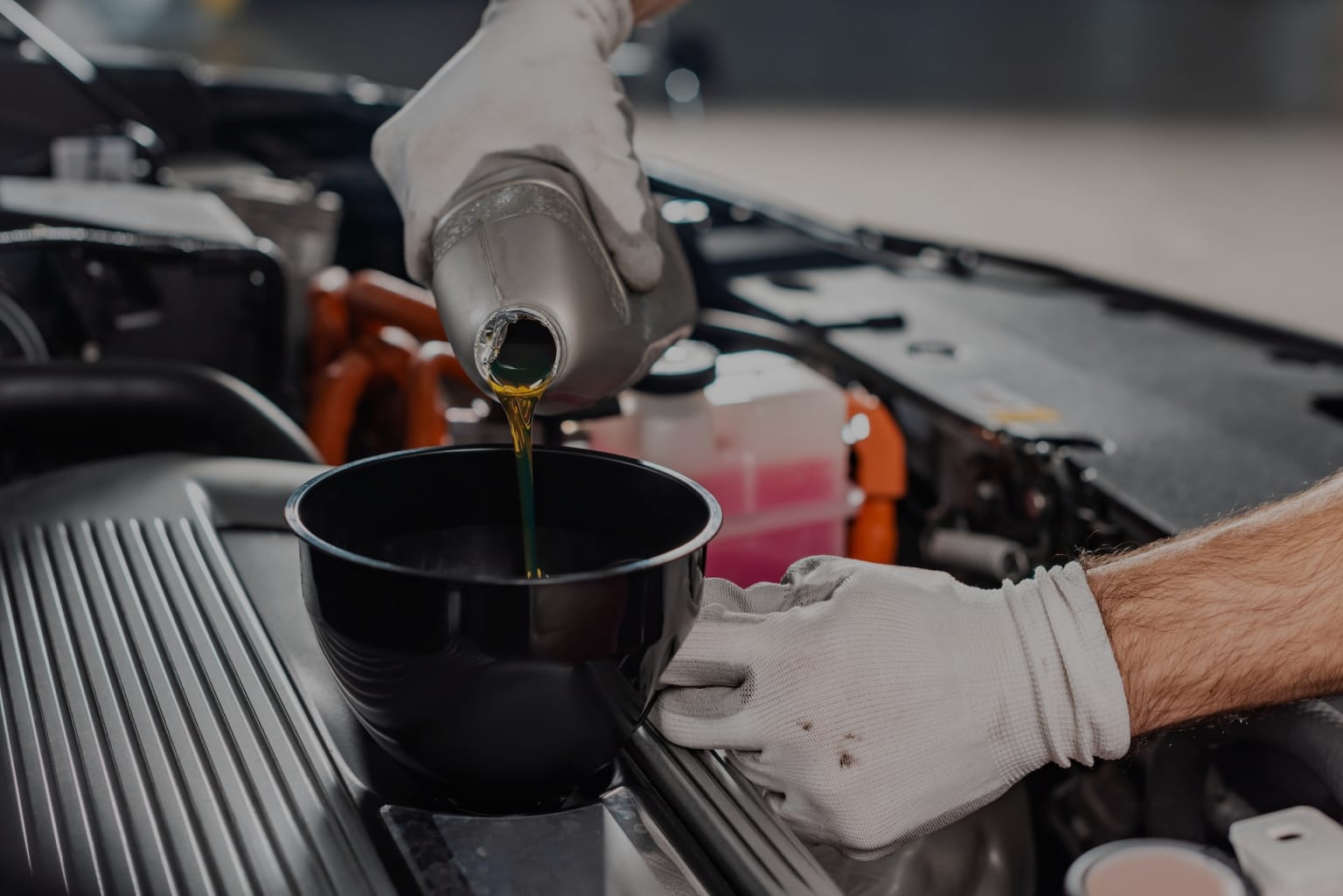 Gloved hands pouring oil into a black bowl over a car engine in a garage.