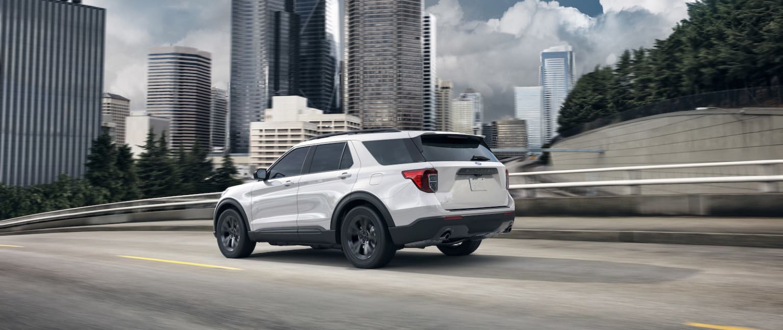 a white suv driving down a city street in front of a tall cityscape with skyscrapers in the background