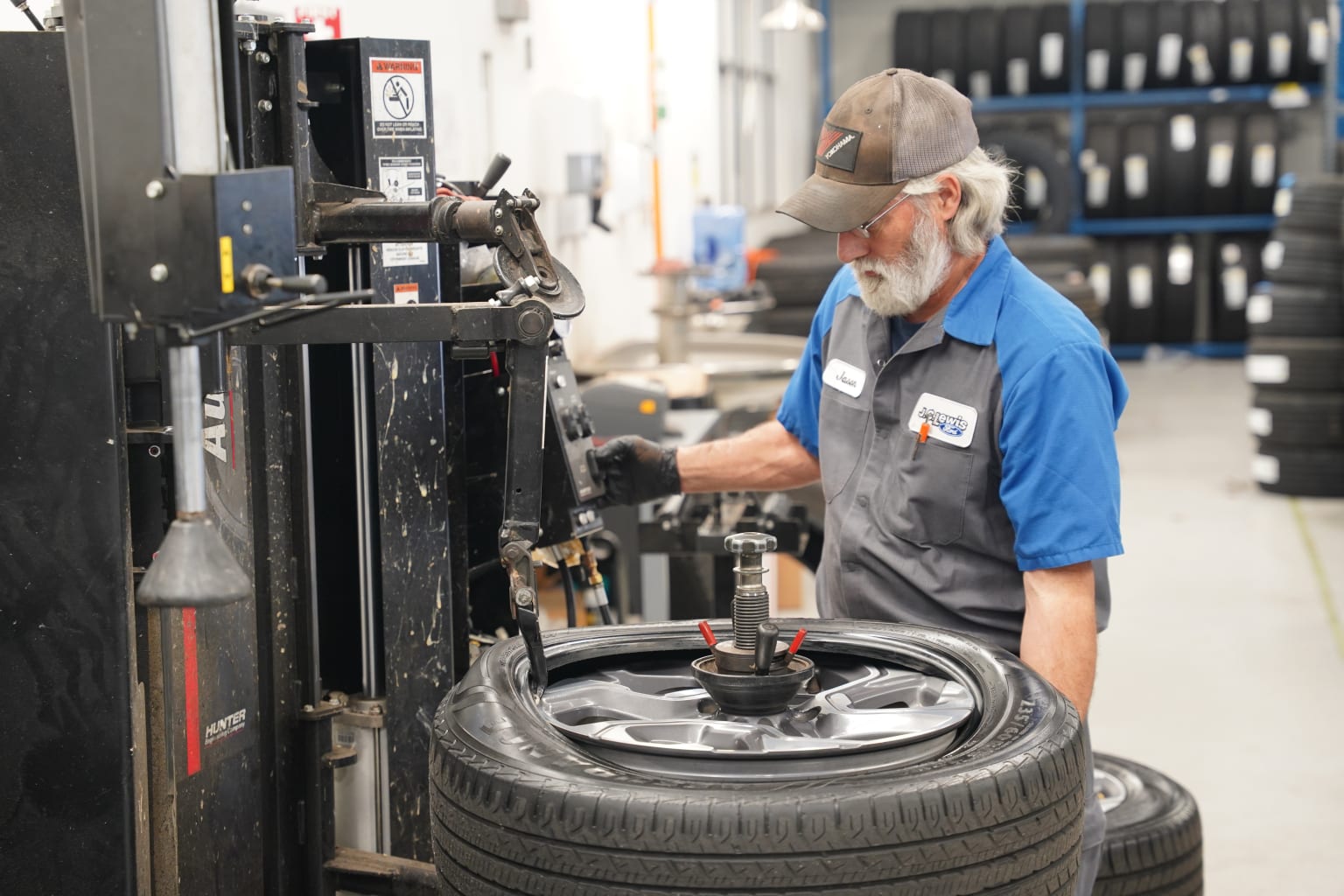 A mechanic in a blue uniform and mask is working on a tire mounted on a machine in a workshop or garage setting, with various tools and equipment visible in the background.