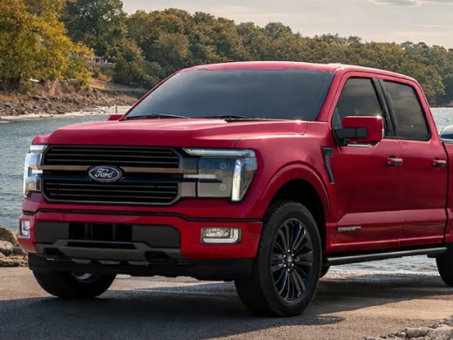 A red truck is parked on a paved surface with a body of water and trees in the background.