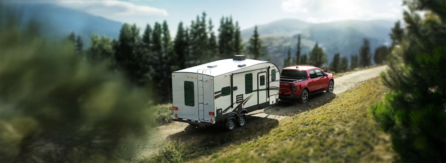 Recreational vehicle being towed by a red pickup truck on a dirt road with green trees and mountains in the background.