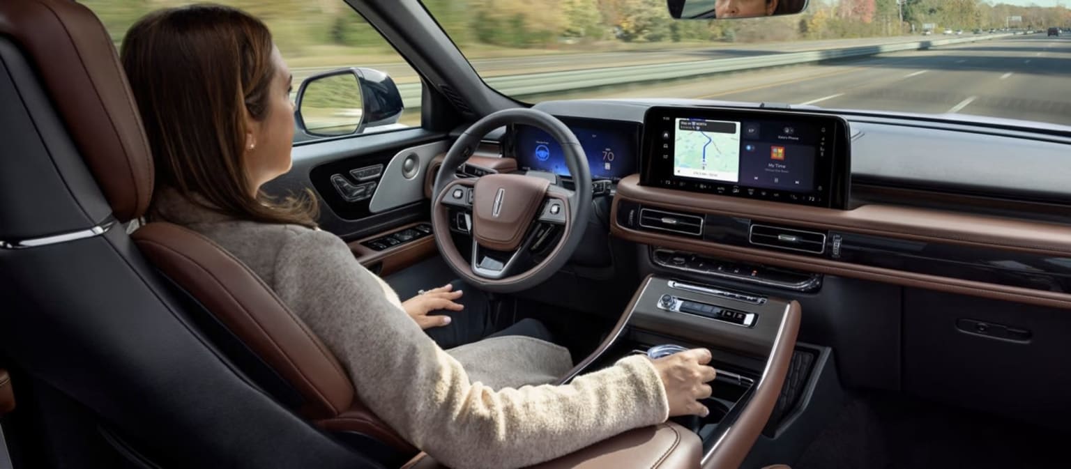 The image shows the interior of a vehicle, with a person sitting in the driver's seat and a large touchscreen display visible on the dashboard. The background outside the vehicle appears to be a road or highway.