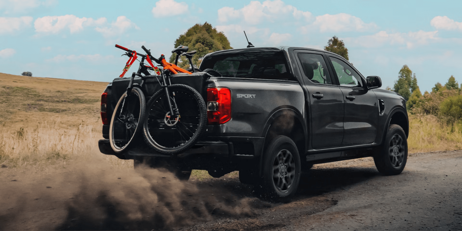 A dark gray pickup truck with two bicycles in the bed kicks up dust on a dirt road with a grassy hill and trees in the background.