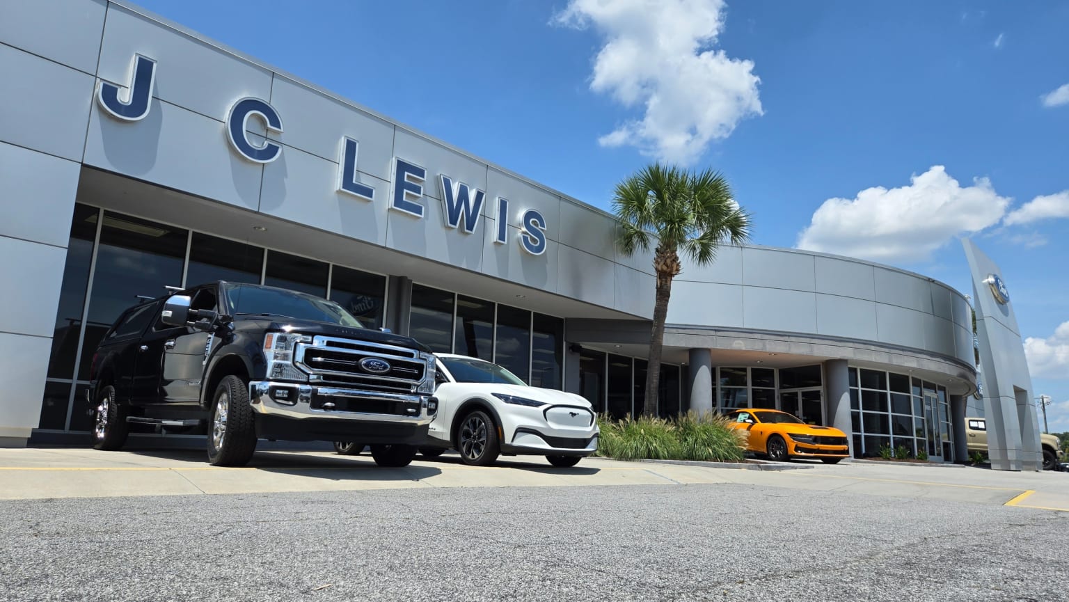 Black truck and white SUV parked in front of a dealership with a yellow sports car visible to the right, under a bright blue sky with scattered clouds.