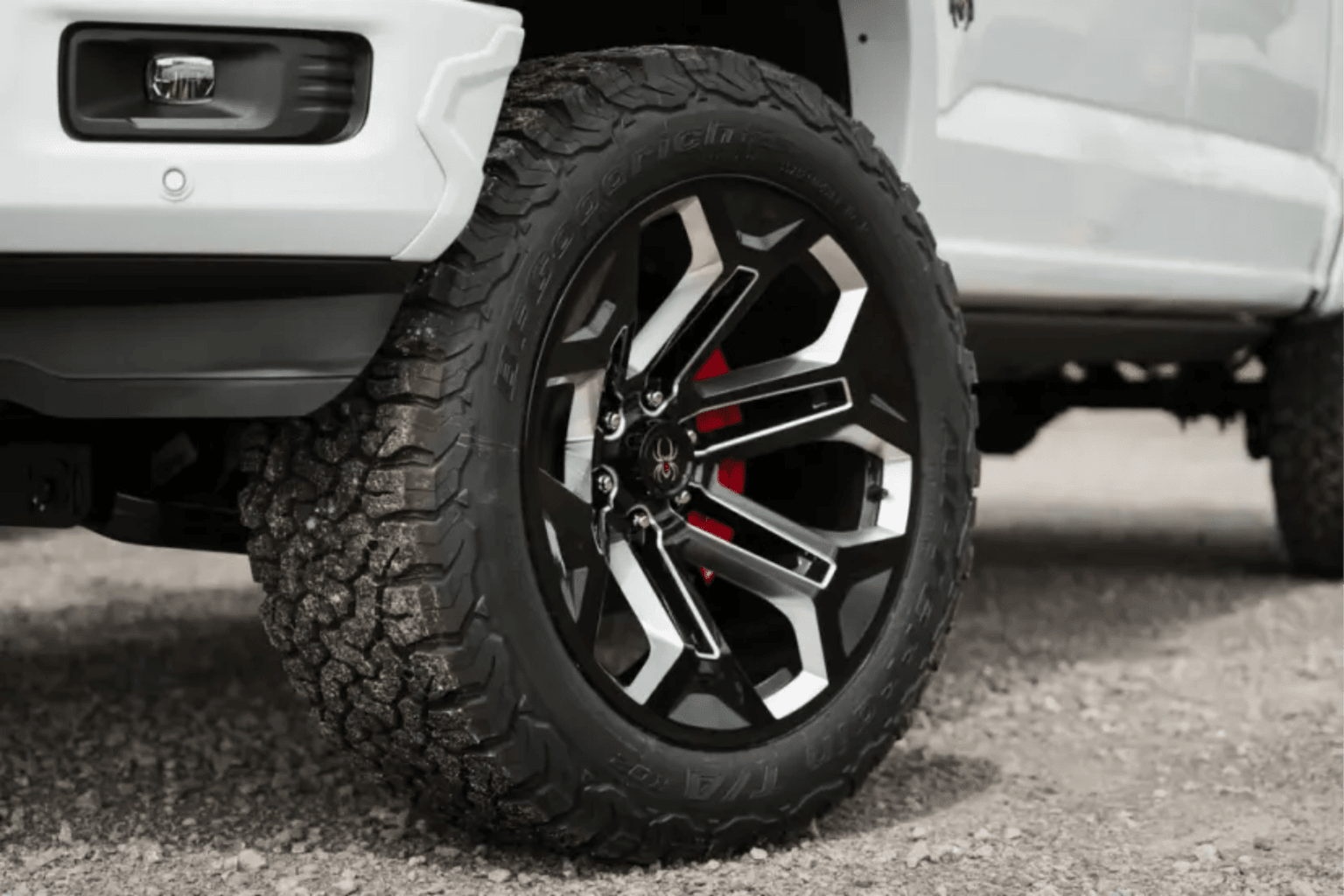 Close-up of an off-road tire and custom rim on a white truck parked on a gravel surface.