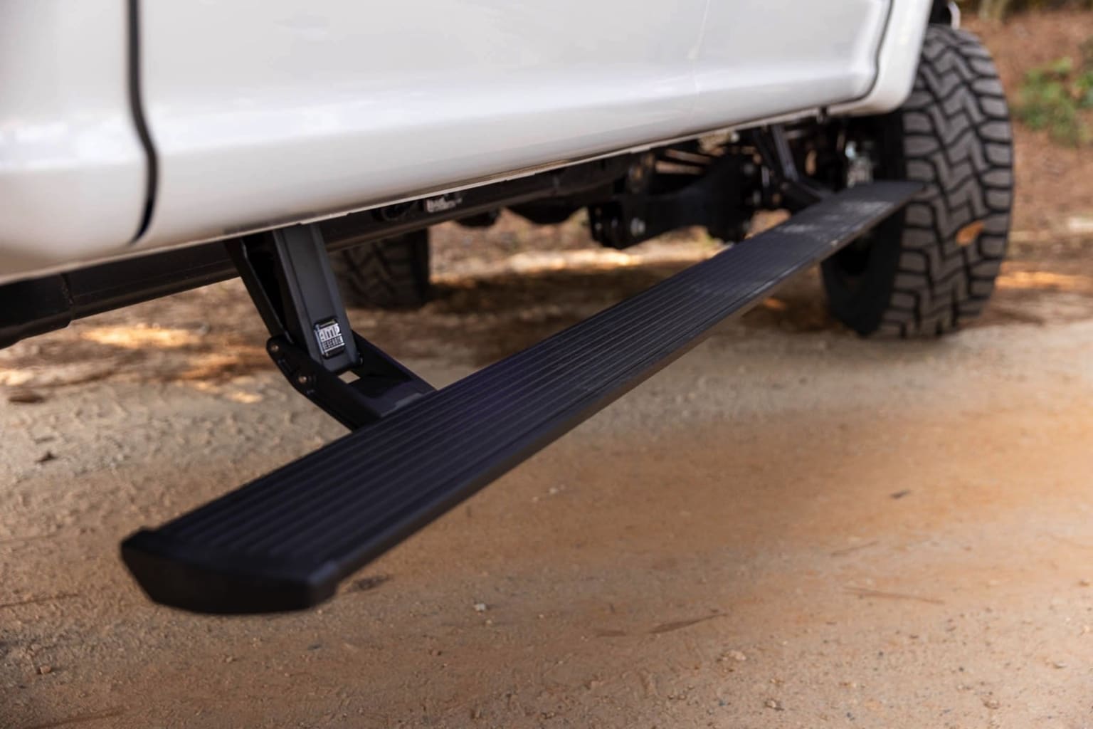 The image displays a side view of a vehicle with a black running board or step attached underneath for easy access. The background shows a dirt or gravel surface, indicating an outdoor or off-road setting.