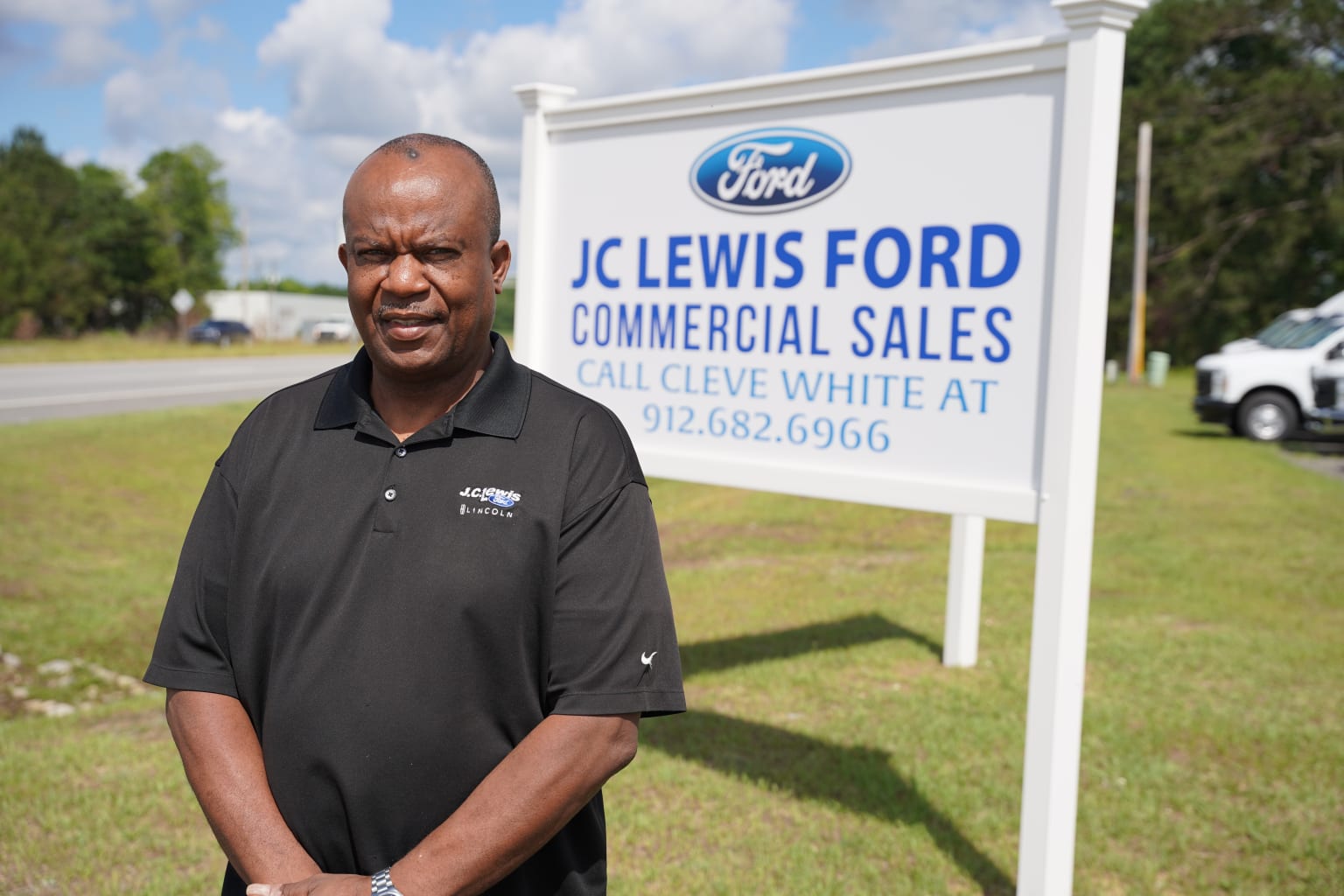 The image shows a man standing in front of a commercial sales sign, with a grassy field and cloudy sky in the background.