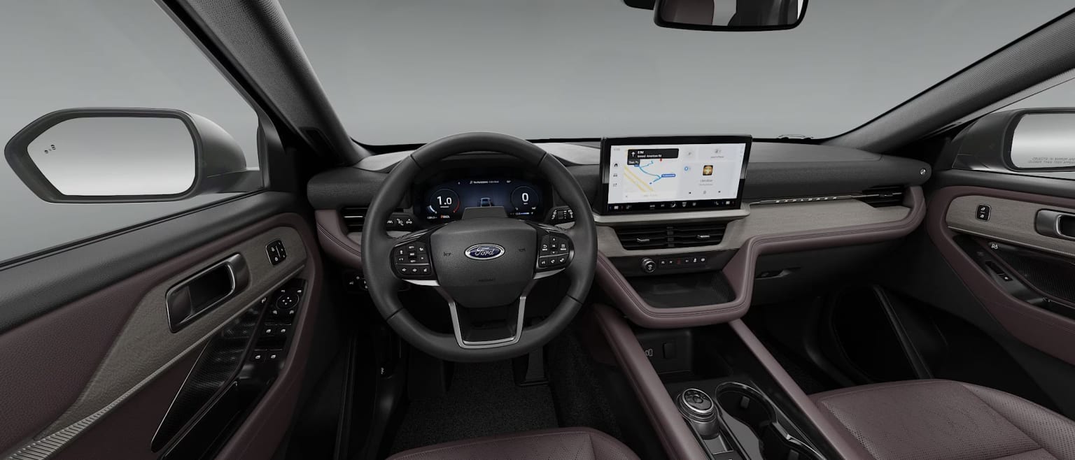 Interior of a vehicle with a steering wheel, dashboard, and a large touchscreen display in the center console, against a neutral gray background.