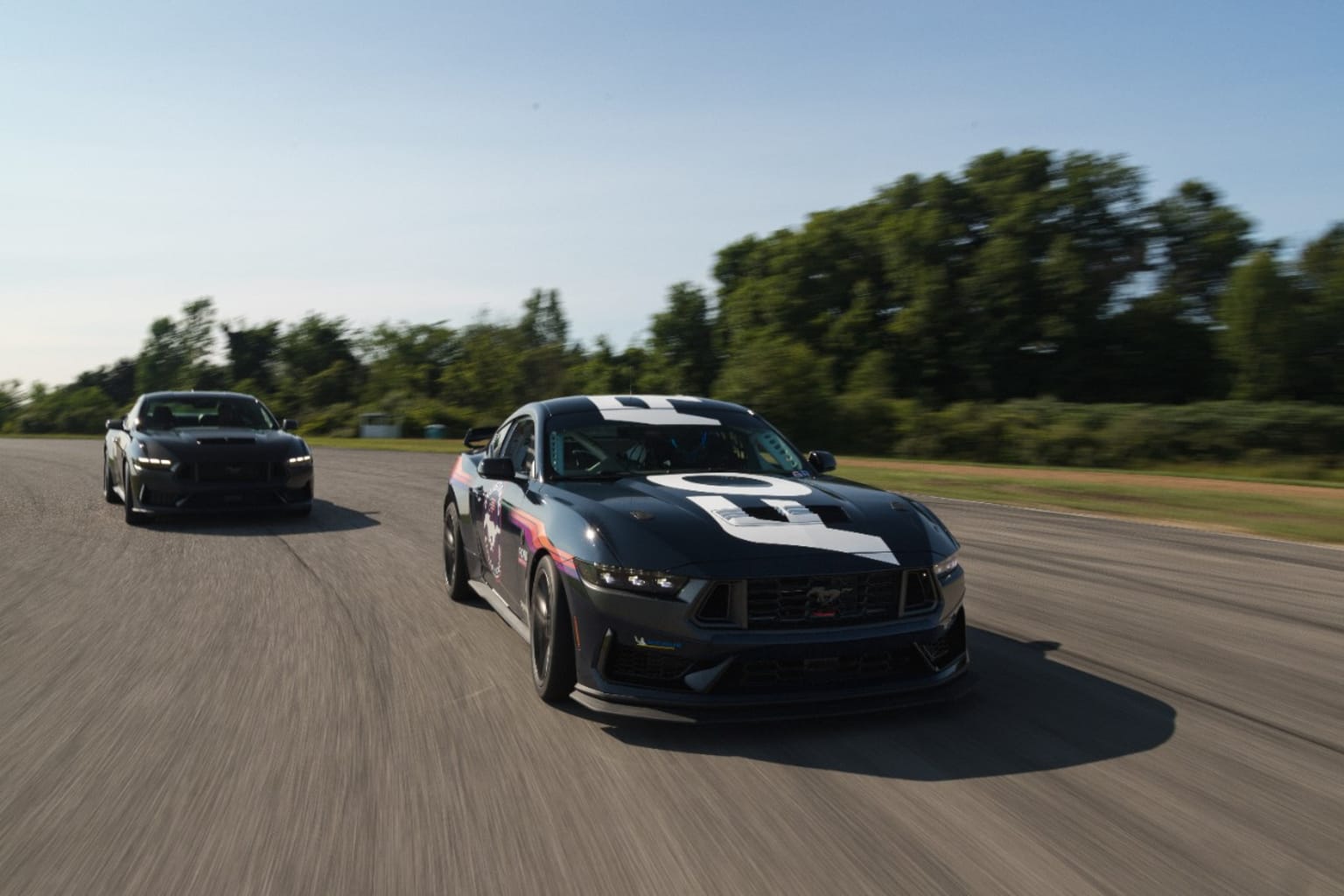 a couple of cars driving down a road next to each other on a race track with trees in the background